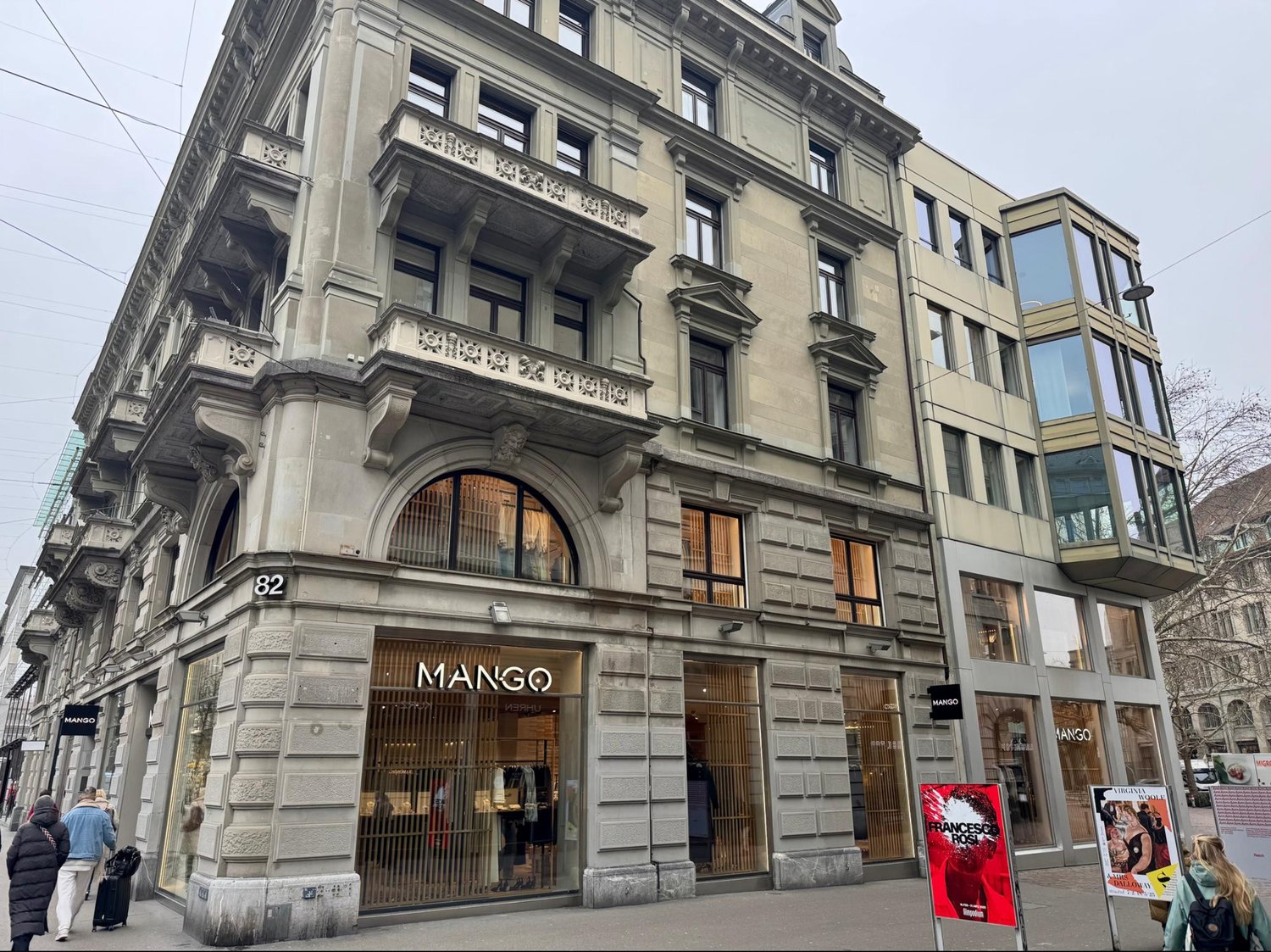 A multi-story building with a classical architectural style, featuring ornate stone facades, arched windows, and decorative elements. The ground floor houses a Mango clothing store, with a large arched entrance. The building appears to be located in an ur