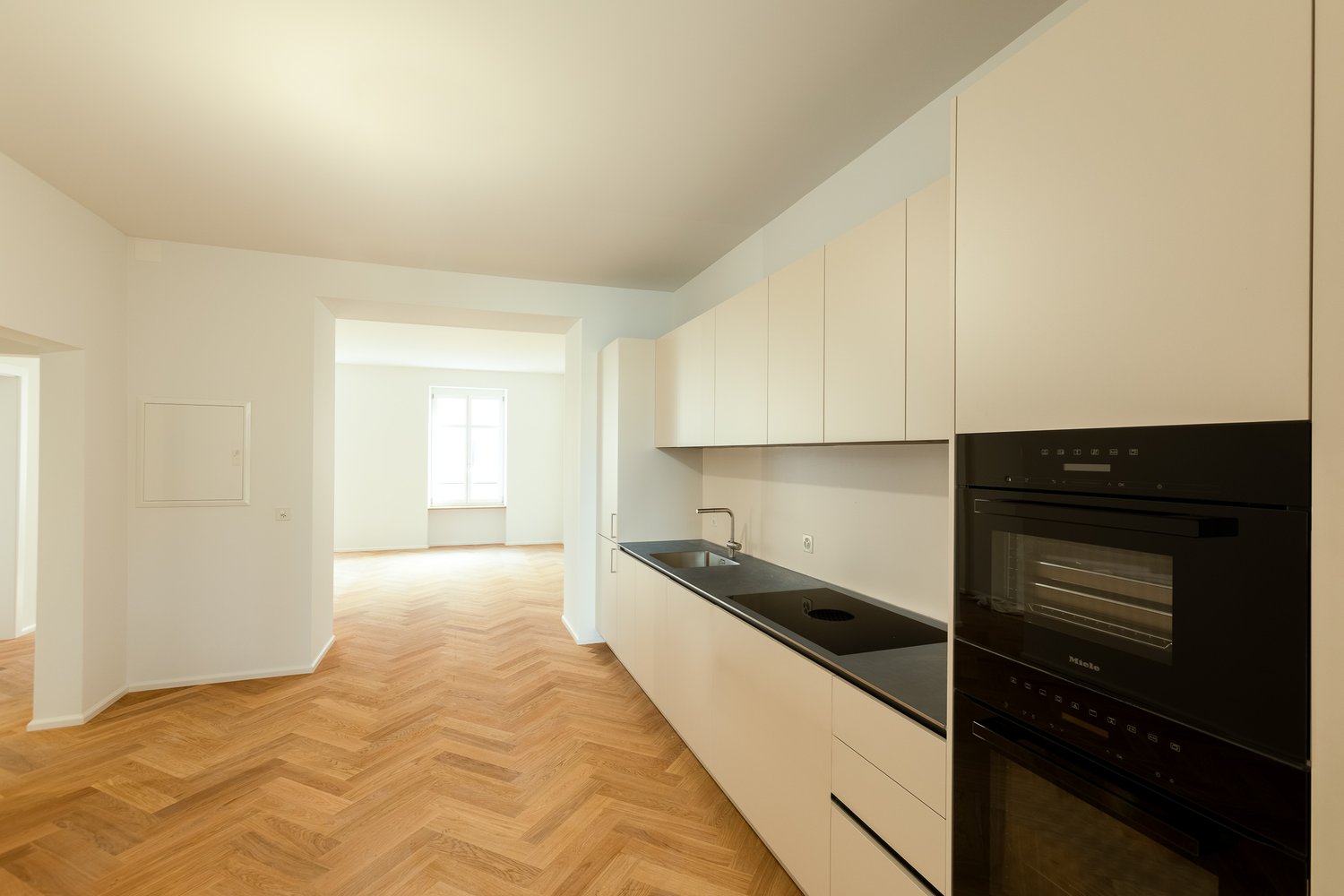 An empty kitchen with white cabinets, wooden flooring, a double sink, and black countertops.