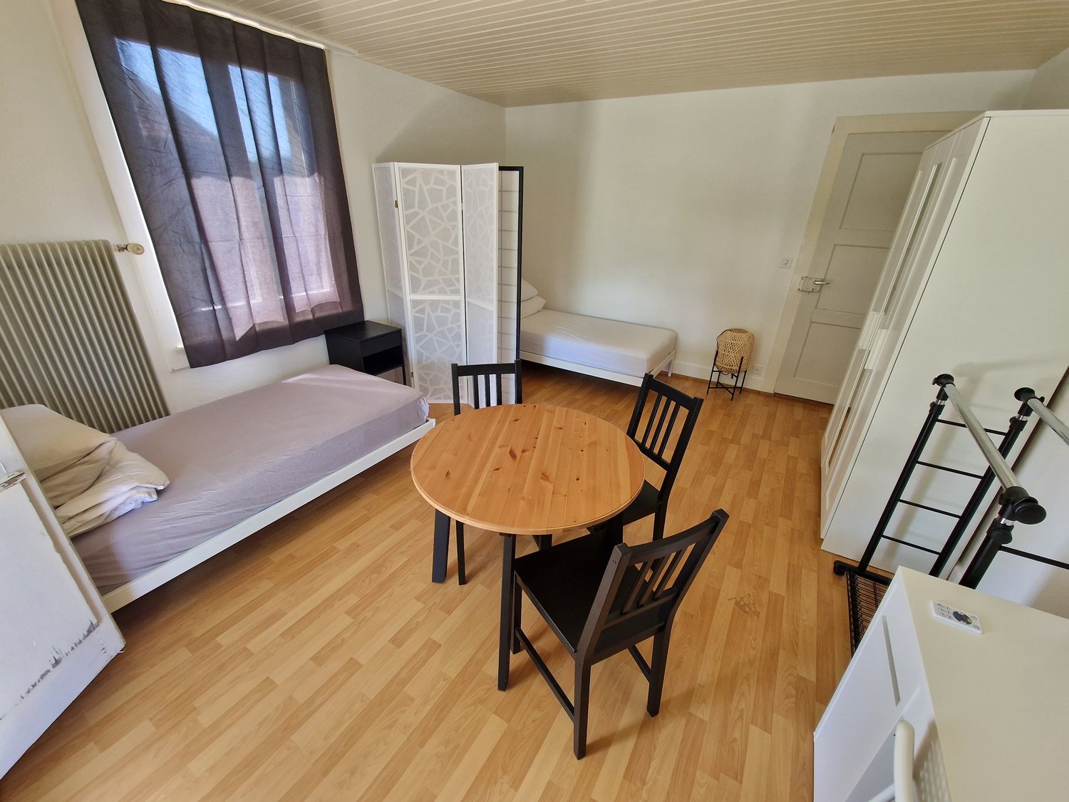 Small room with wooden floor, single bed, round table, 2 chairs, wardrobe, heater