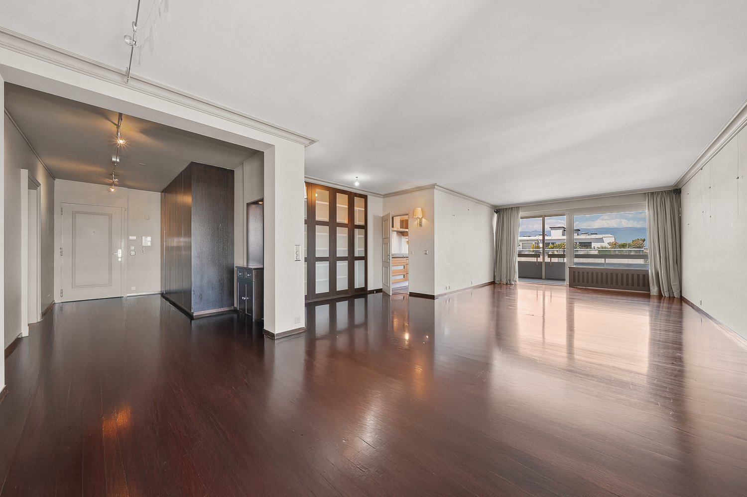 Spacious open-concept living area with hardwood floors, large windows, and a balcony. The room features a modern design with a built-in shelving unit dividing the space. The apartment appears to have a kitchen and other rooms beyond the living area.
