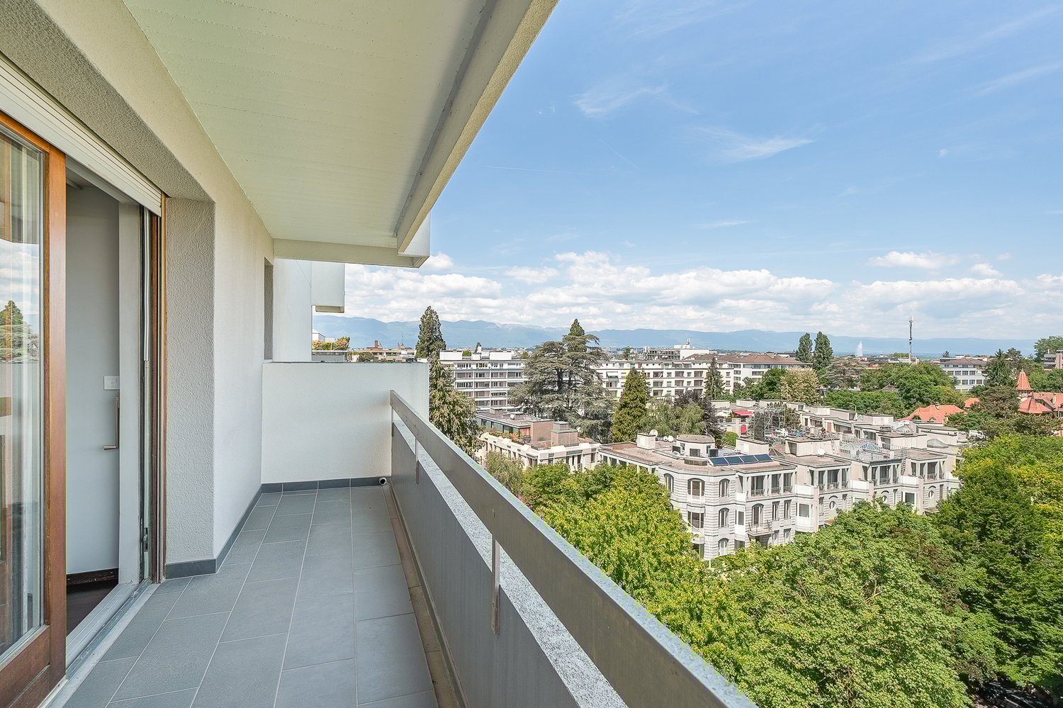 Spacious balcony with a panoramic view of the city, including residential buildings, trees, and mountains in the distance. The balcony has a tiled floor and is accessible through large windows or sliding doors.