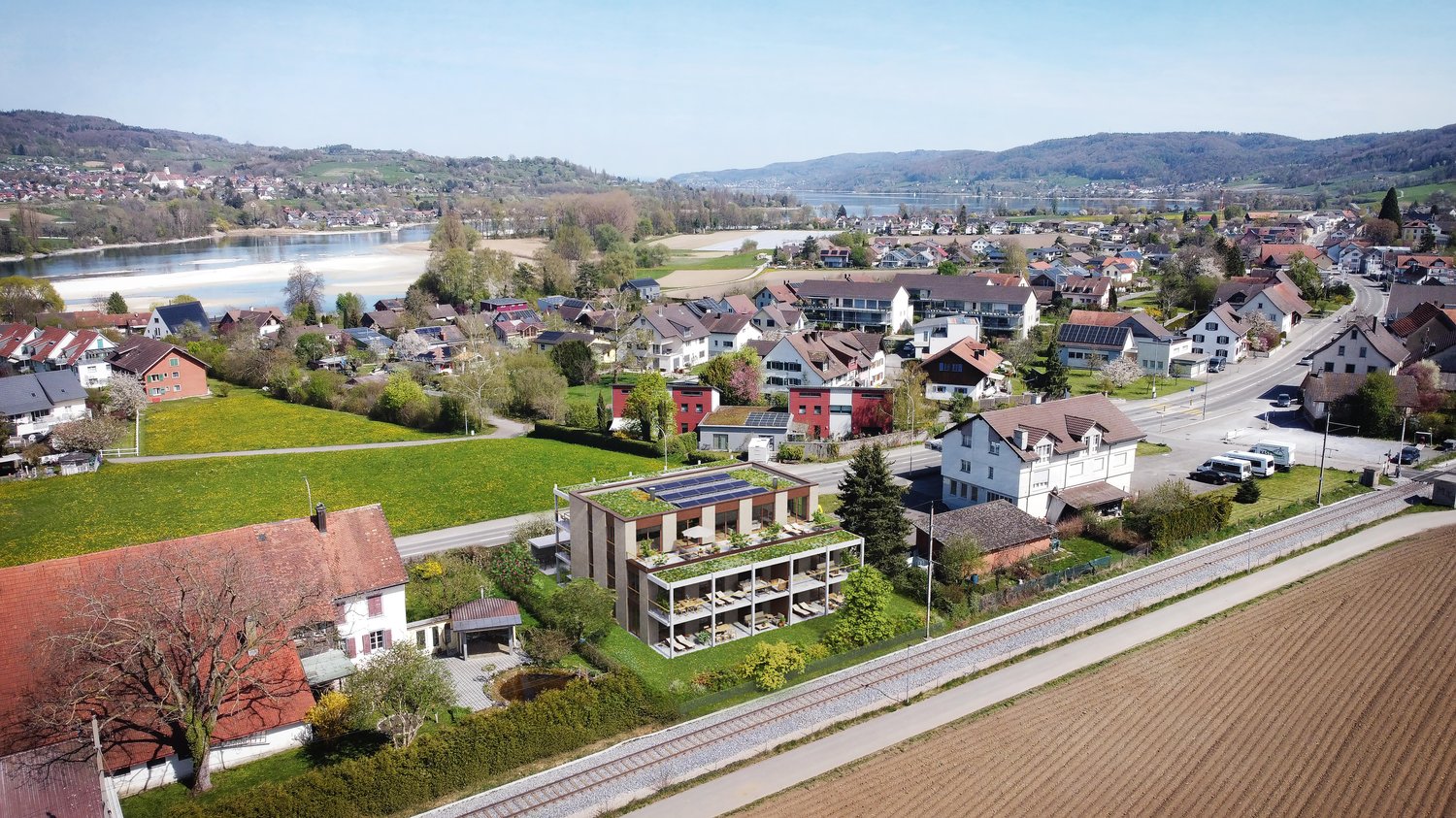 This image shows a picturesque town nestled along a lake or river, with rolling hills and mountains in the background. The town features a mix of residential and commercial buildings, some with green roofs or balconies. There is a road running through the