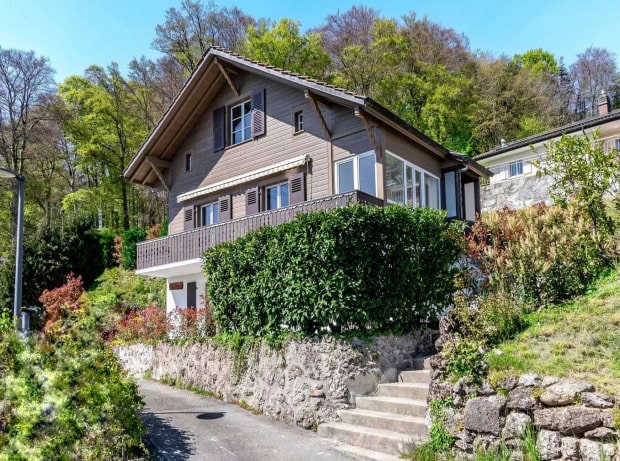 2-story wooden house with a balcony, surrounded by trees and greenery, situated on a hillside with stone steps leading up to the entrance.