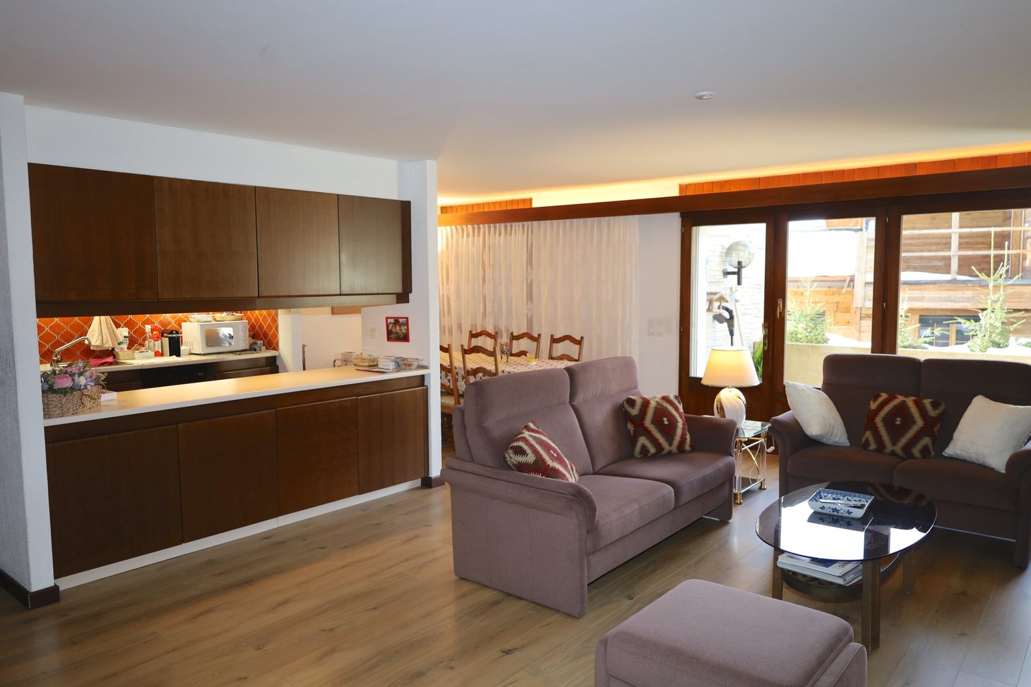 modern kitchen with wooden cabinets, dining area with chairs, two couches, coffee table with books, lamp, large windows