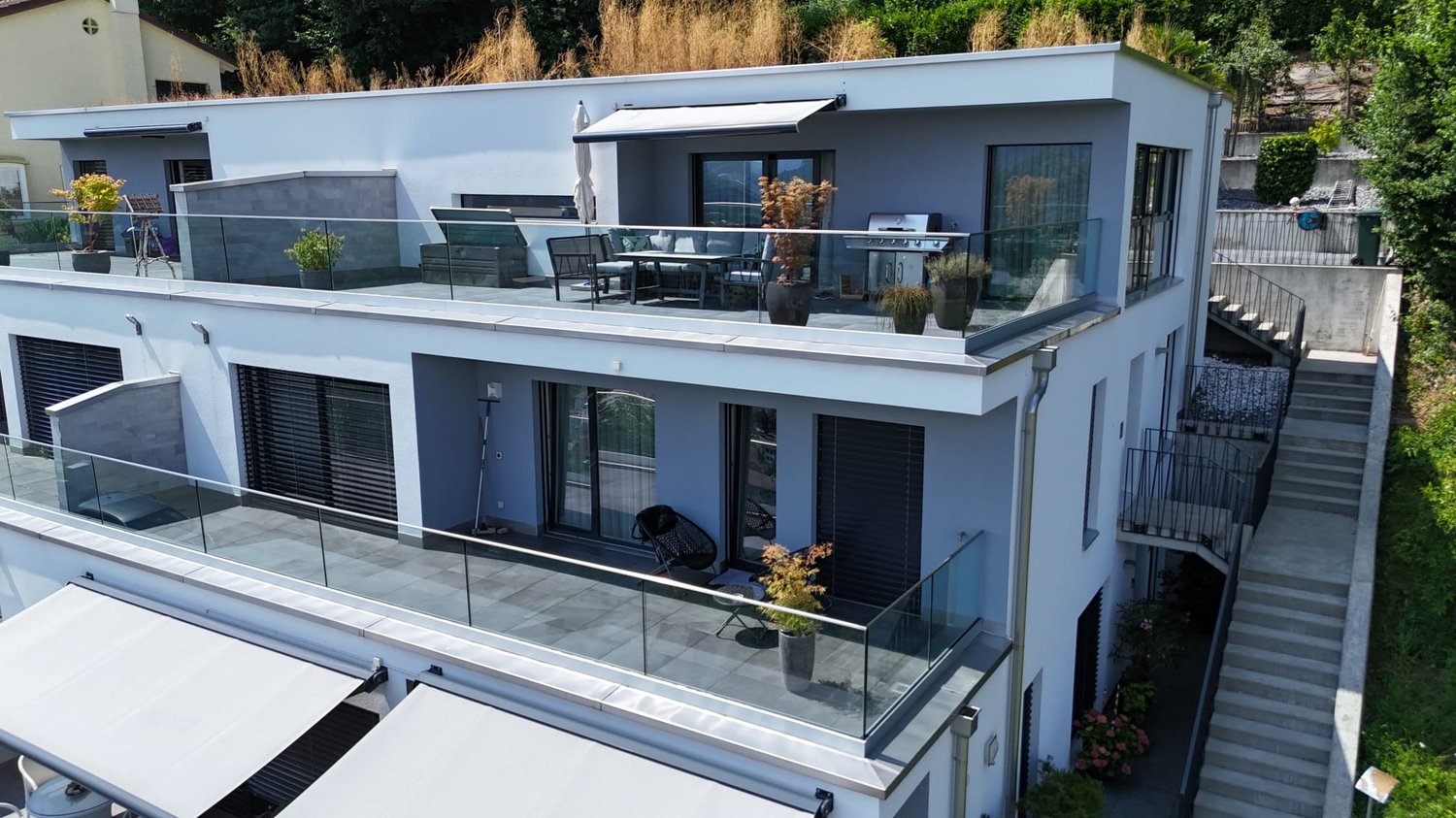 2-story modern house with a flat roof, glass balconies, and a garage. The exterior is painted in a light blue color with large windows. There is a patio area with outdoor furniture on the ground floor.