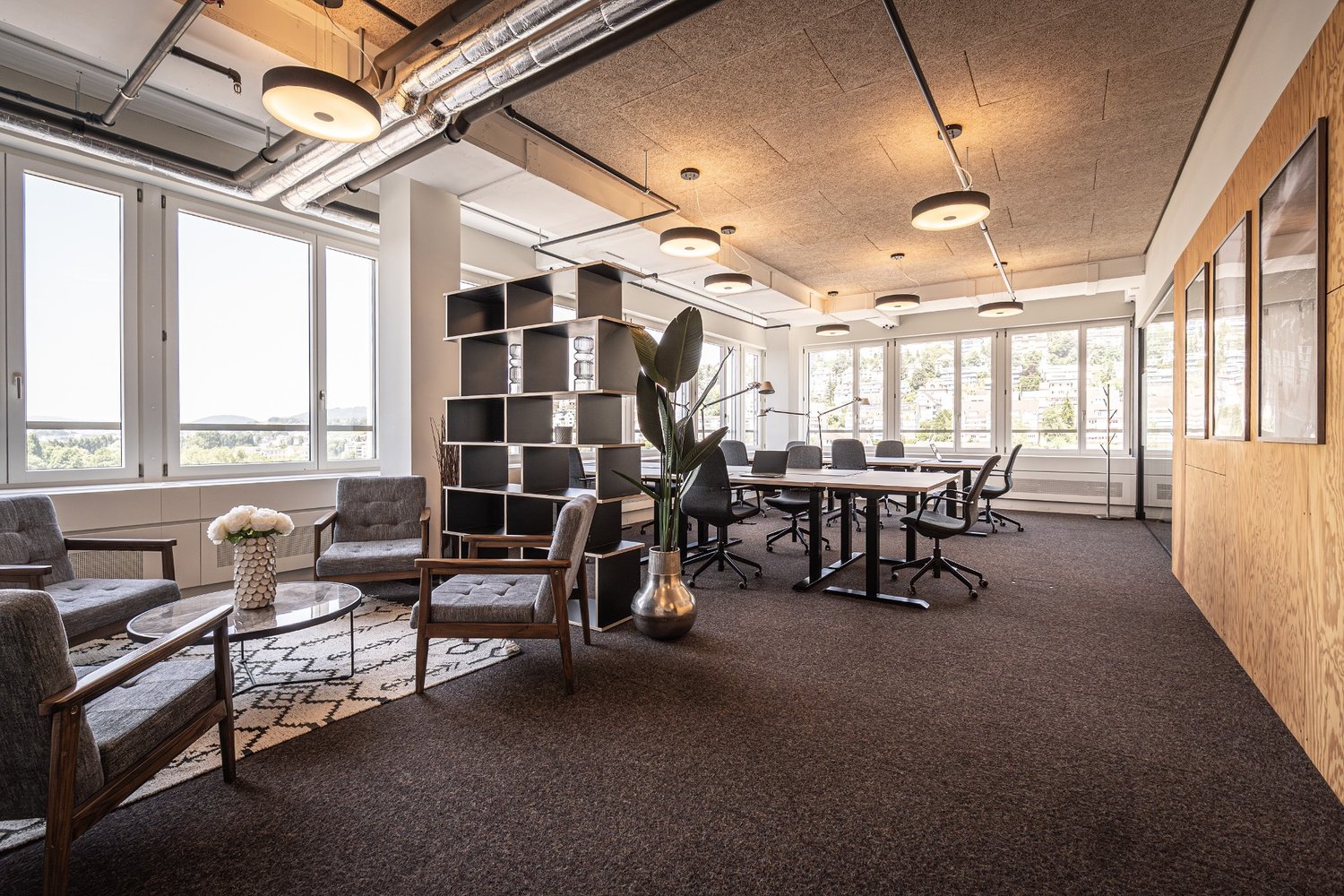 Modern office room with open layout, carpeted floor, gray chairs, tables, glass windows, pendant lighting, and greenery