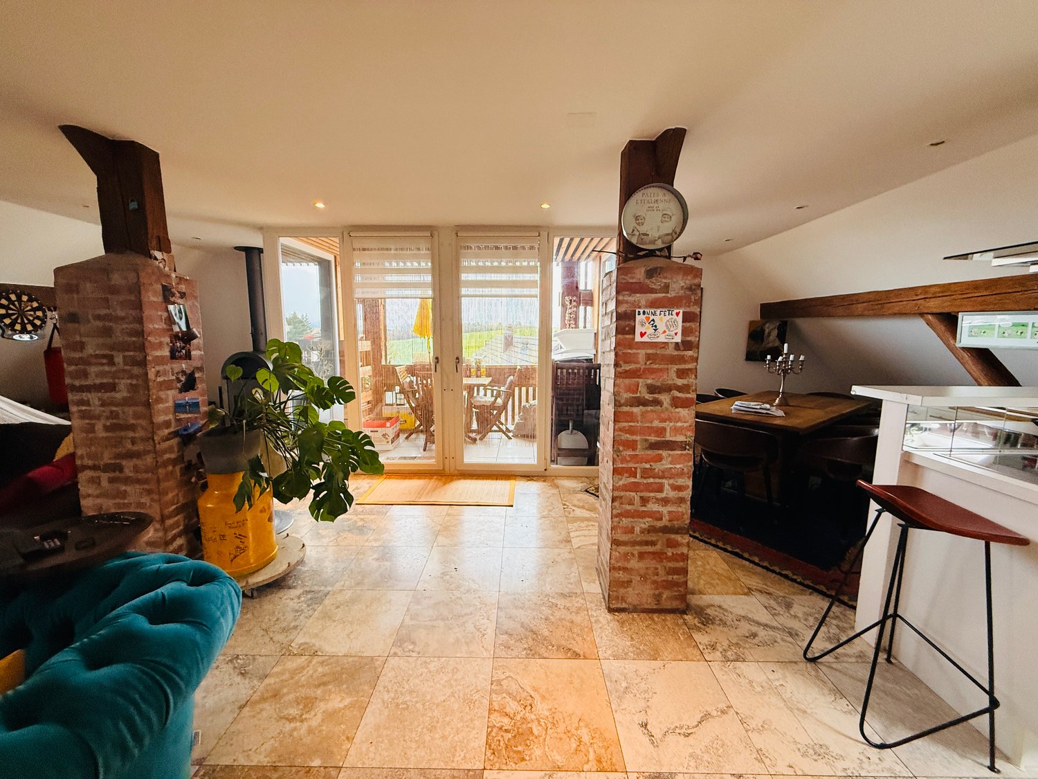 Open floor plan, brick pillars, kitchen island, bar stools, sofa, large floored window, balcony, fireplace, potted plant, table, chairs