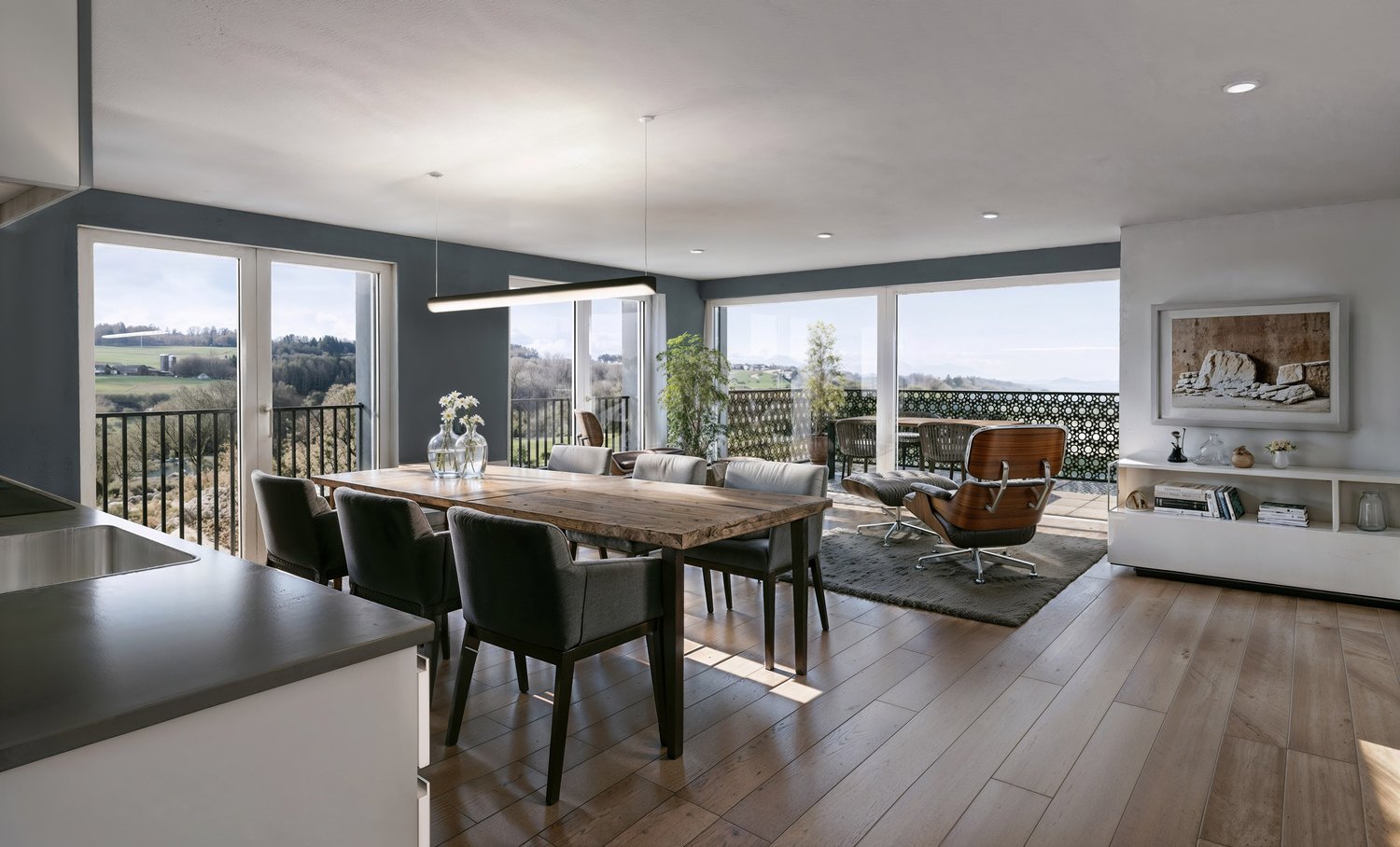 Open concept living room and dining area with large windows overlooking a balcony or terrace, hardwood floors, modern furniture including a wooden dining table and leather chairs, and a cozy seating area with a leather armchair.