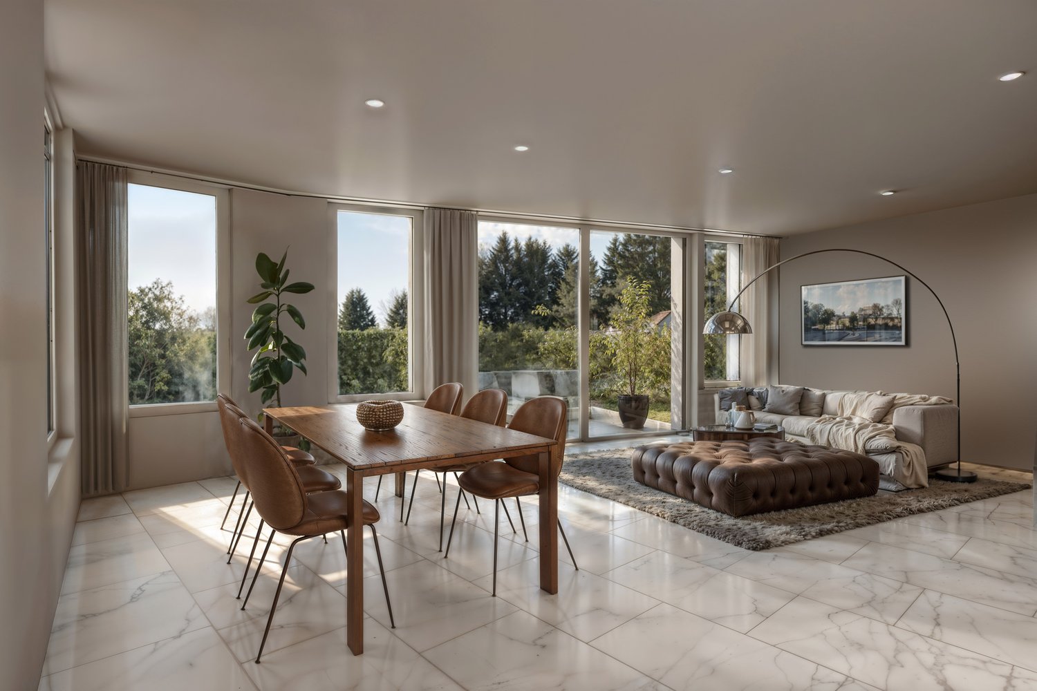 Spacious open-concept living and dining area with large windows offering a scenic view of the surrounding trees and nature. The living room features a comfortable tufted leather sofa and a large floor lamp. The dining area has a wooden table with leather