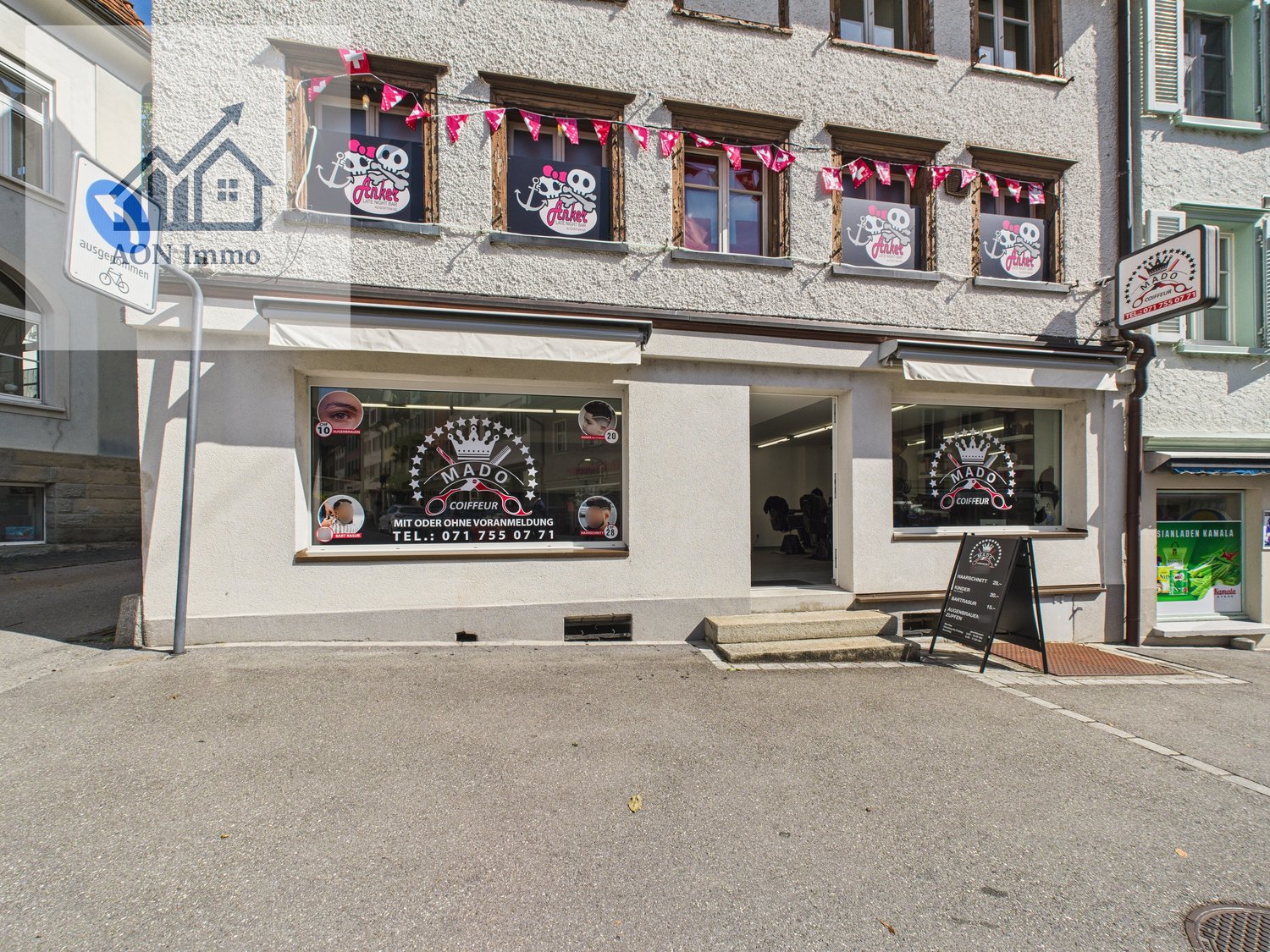two-story commercial building with white walls, multiple windows, and signage indicating a salon named MADOK. There is a blackboard sign, decorative flags, and some advertisement in the windows