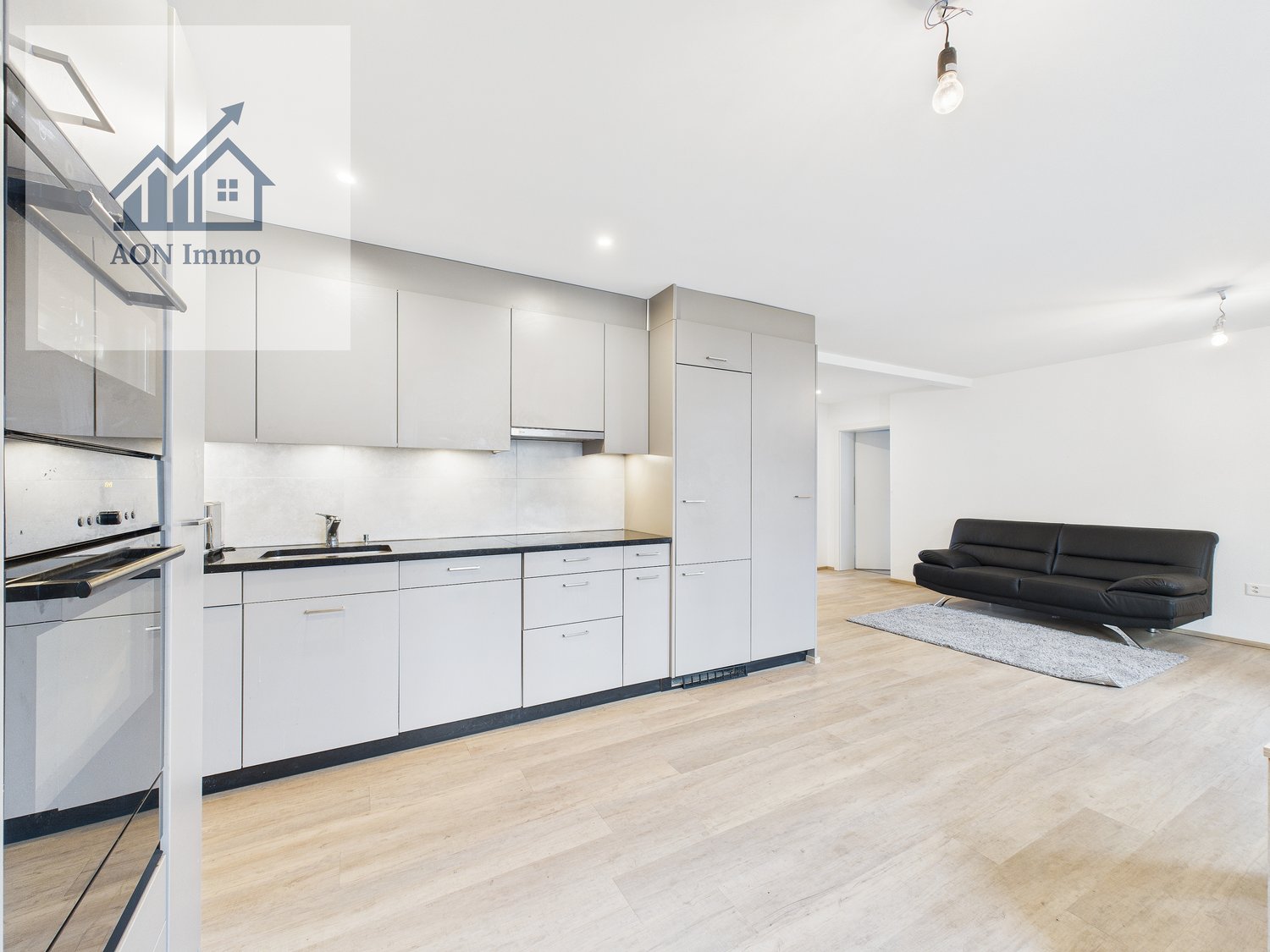 Modern kitchen with grey cabinets, white appliances, wooden flooring, open living room area with black sofa on a rug