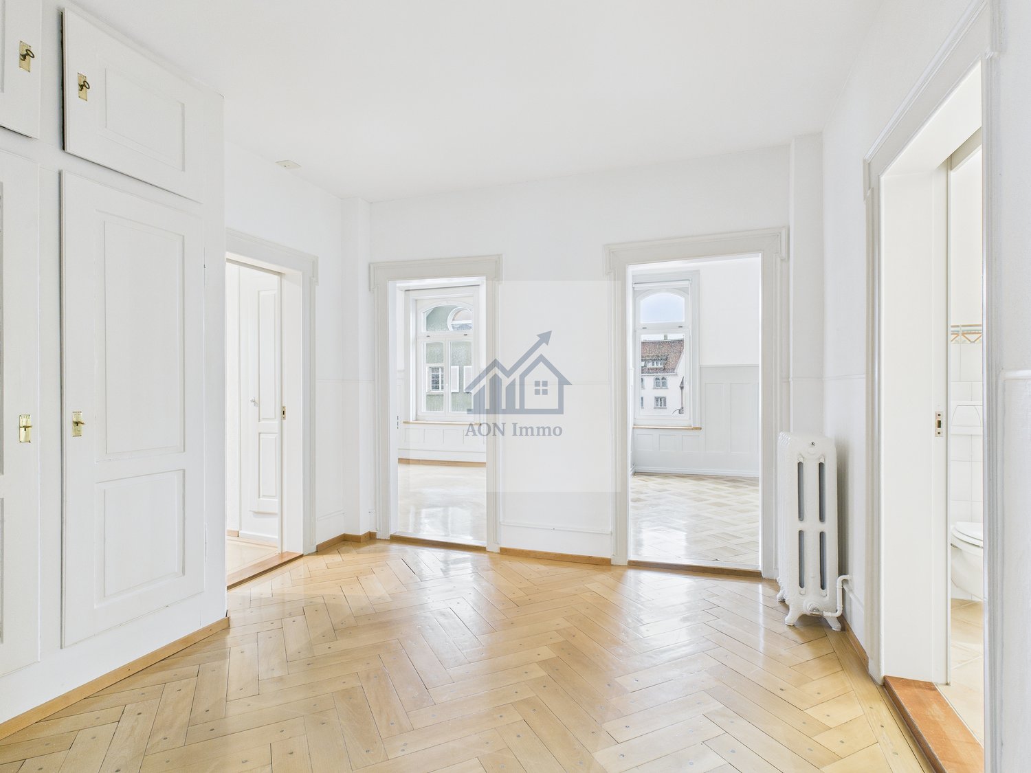 A spacious and well-lit room with white walls, wooden floors, and multiple windows allowing natural light to enter. The room appears to be clean and empty, with visible doors leading to other areas. A radiator is visible on the wall.