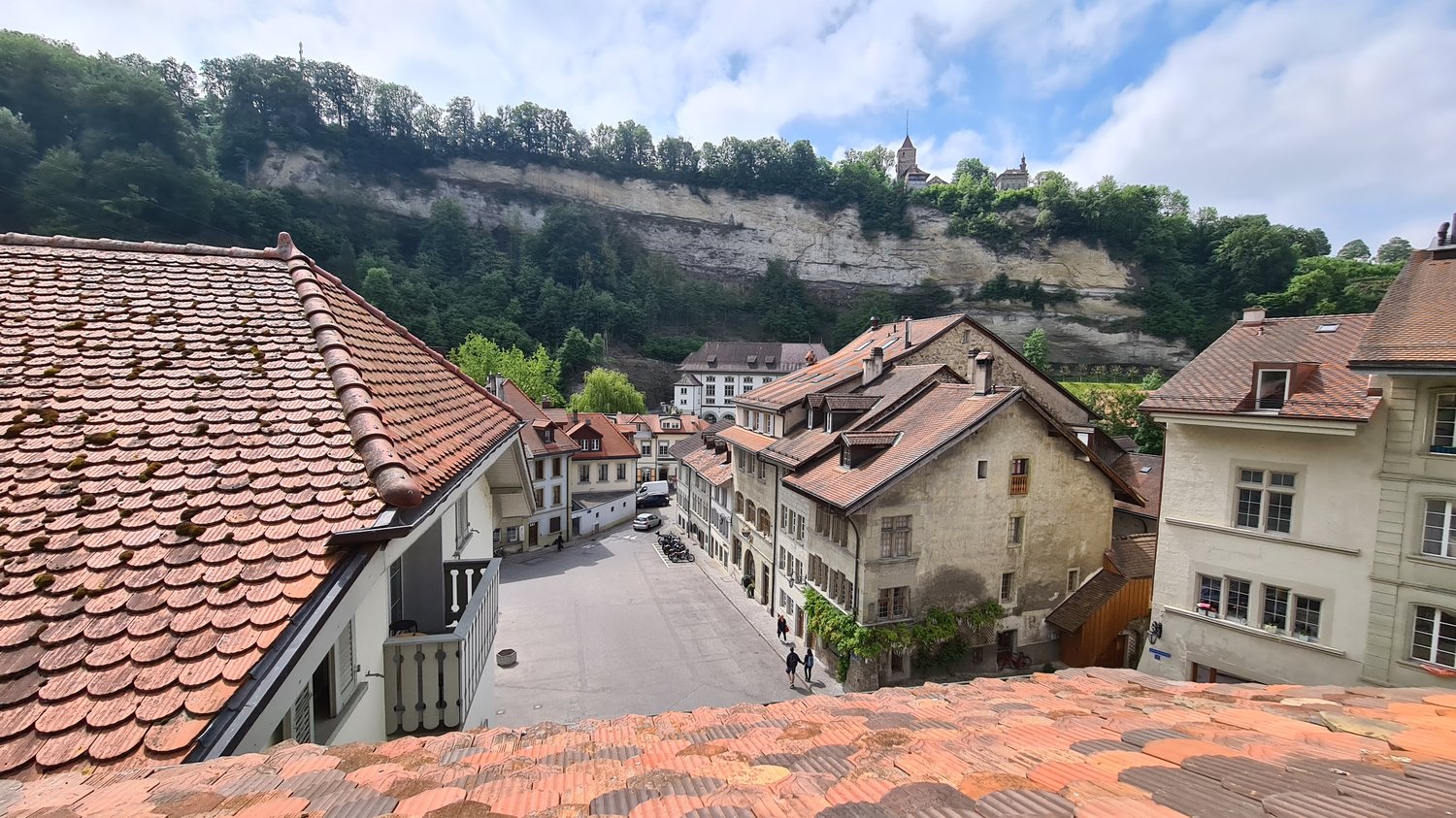 stone wall, terracotta roof, old building, hill in the background, cars on street, group of people walking, clear sky with clouds