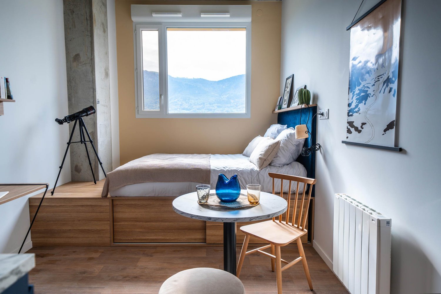 Cozy bedroom with large window offering a scenic mountain view, wooden platform bed, side table with decorative items, and a telescope for enjoying the outdoor scenery.