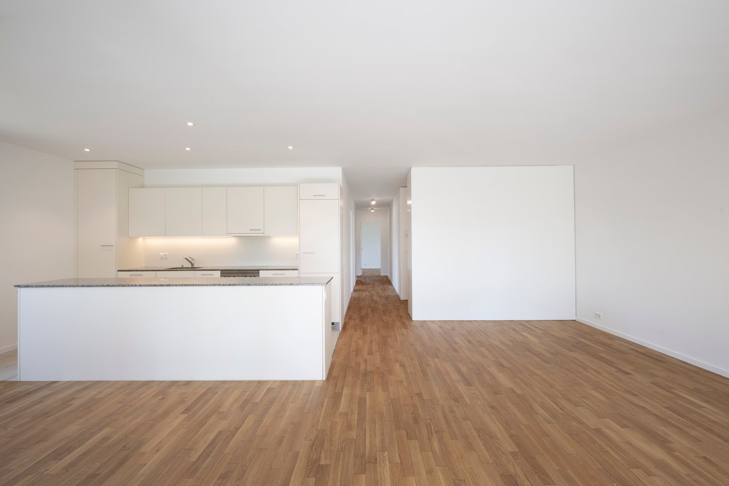 Empty room with white walls, hardwood floors, island counter in kitchen area, white cabinets, open hallway.
