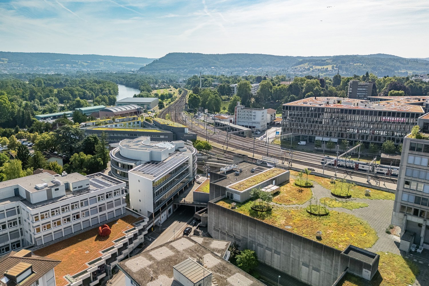 Aerial view of a city with a mix of modern and older buildings, surrounded by hills and greenery. The image shows a train station, parking areas, and green roofs on some of the buildings.