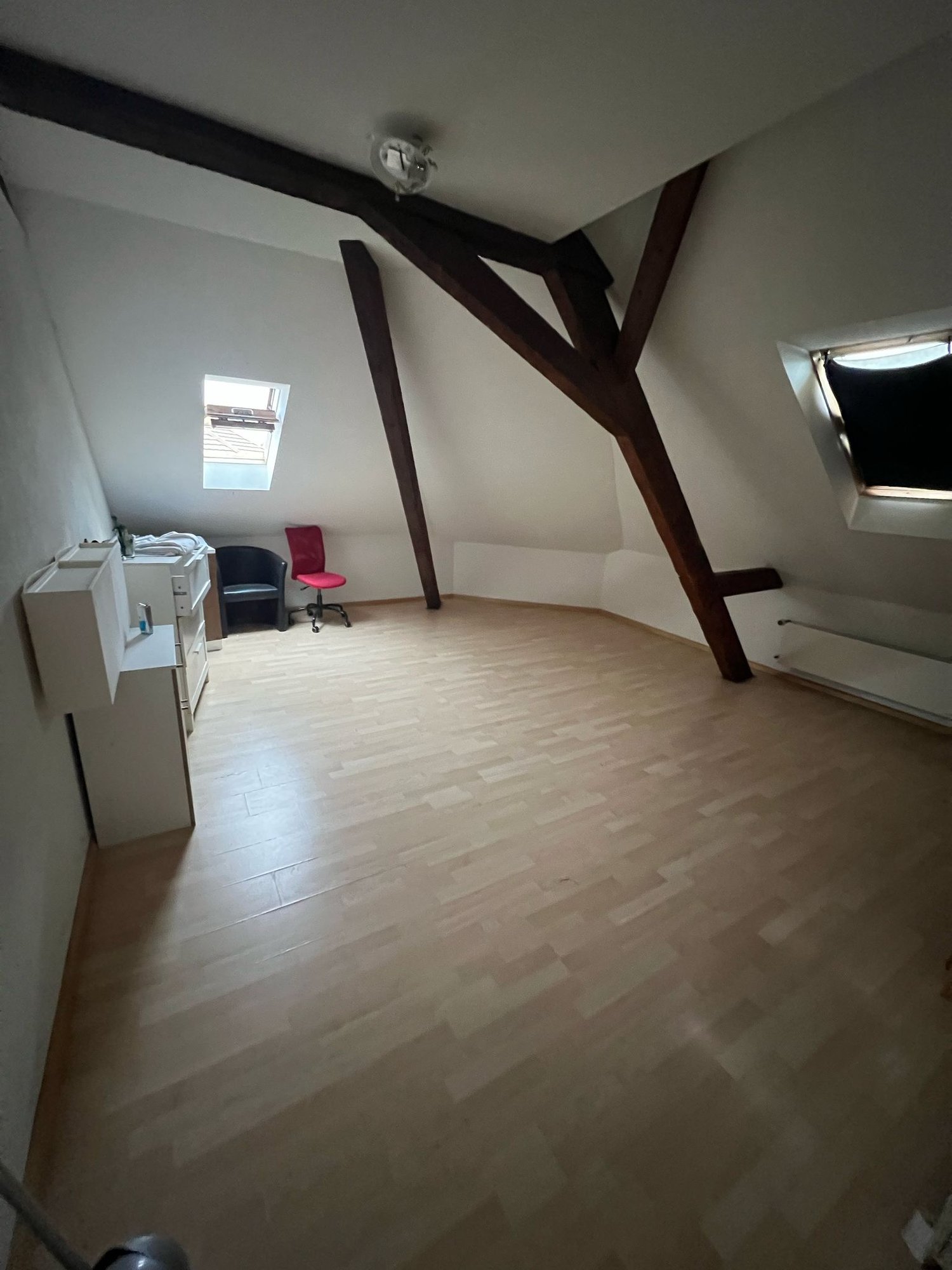 Empty attic room with wooden beams, hardwood floors, two skylight windows, radiator, two chairs, and a small wardrobe with drawers