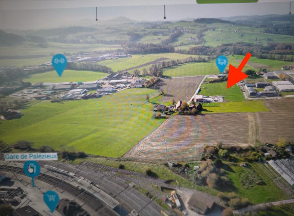Aerial view of a rural area with fields, buildings, and a railway. The highlighted area shows a small house with a red arrow pointing towards it.