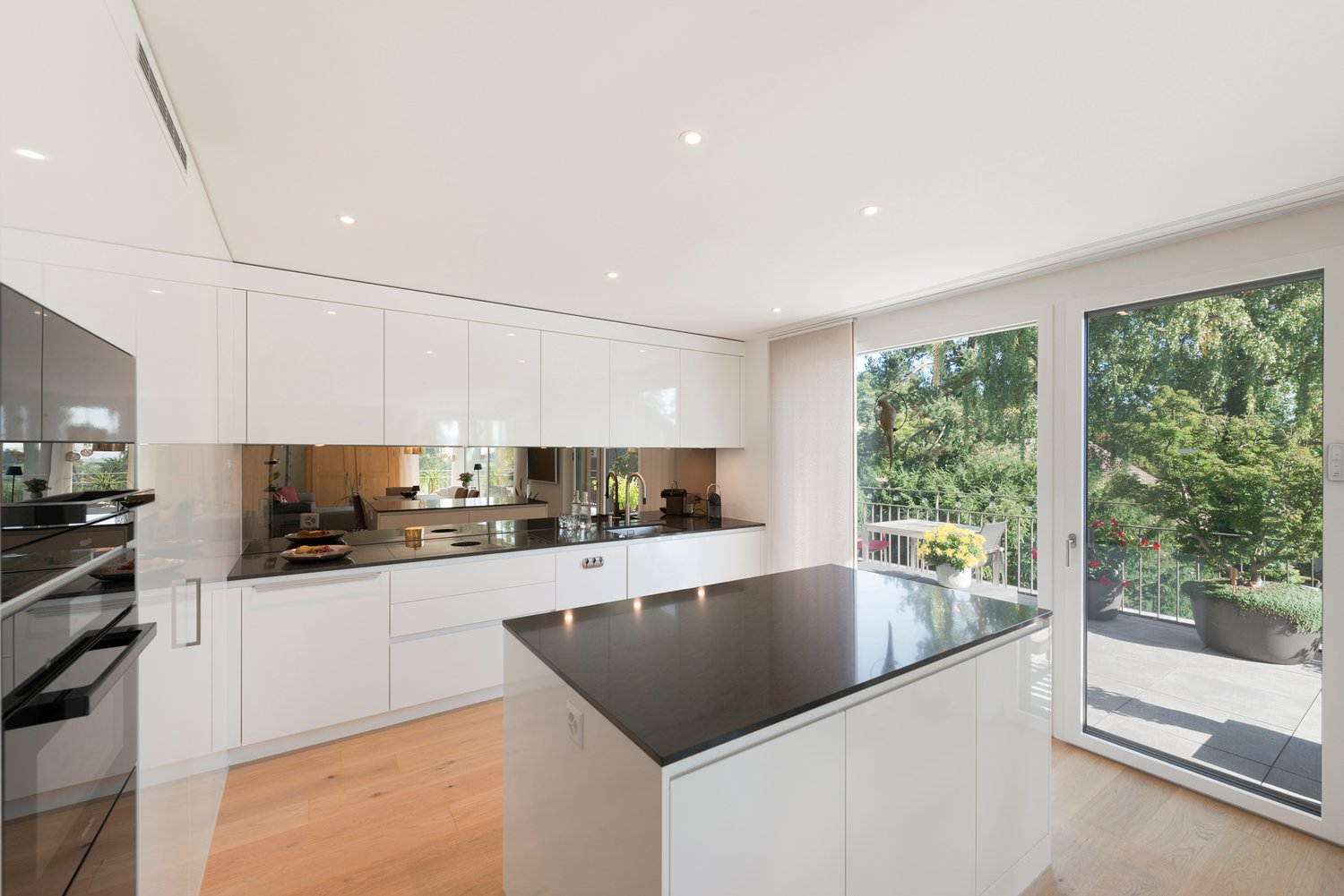 Modern kitchen with white cabinets, island countertop, gas stove, and black granite countertop. It has large windows leading to a balcony with plants and views of the outdoors.