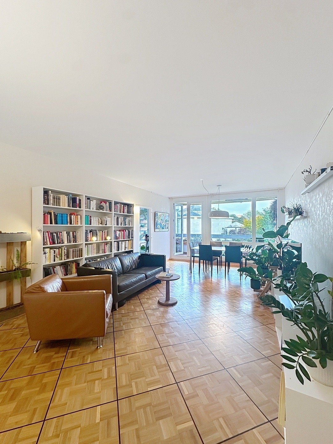 living room with a leather sofa, a leather chair, a side table, a bookcase with books, a round dining table, chairs, potted plants, wooden floor, and windows