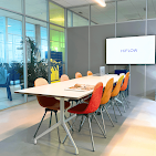 Empty meeting room with glass walls, long white table, orange and blue chairs, screen, whiteboard
