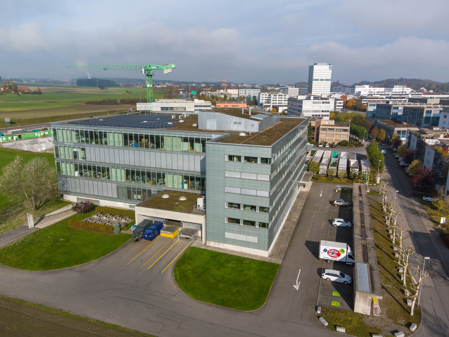 Office building, 4 stories, large windows, grey and white exterior, solar panels on the roof, parking area with cars