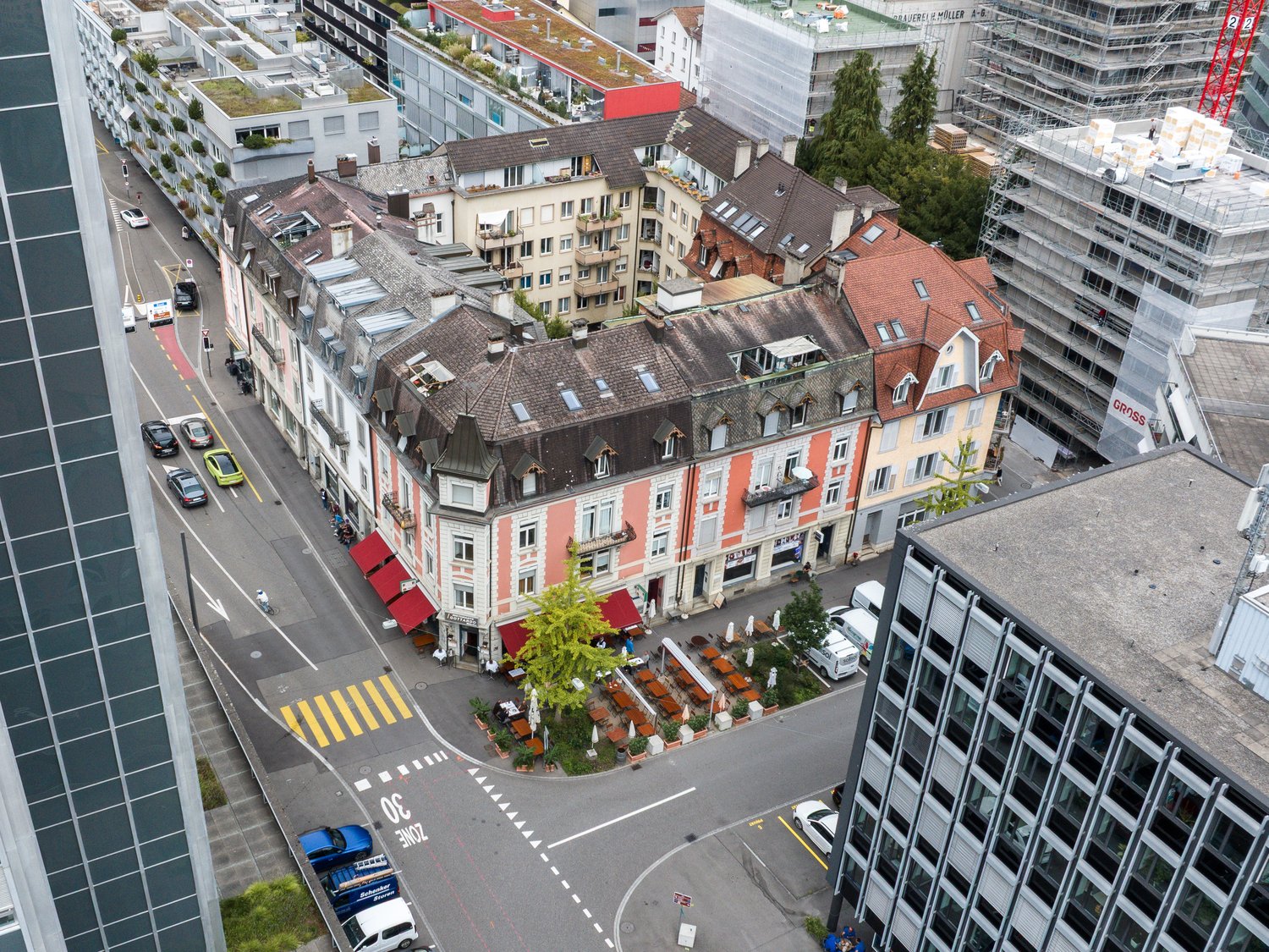 The building is a mixed-use building with a commercial ground floor and residential apartments above. The building is painted yellow with a brown roof. The roof features multiple dormer windows and a chimney. There is a patio with tables and chairs in fro