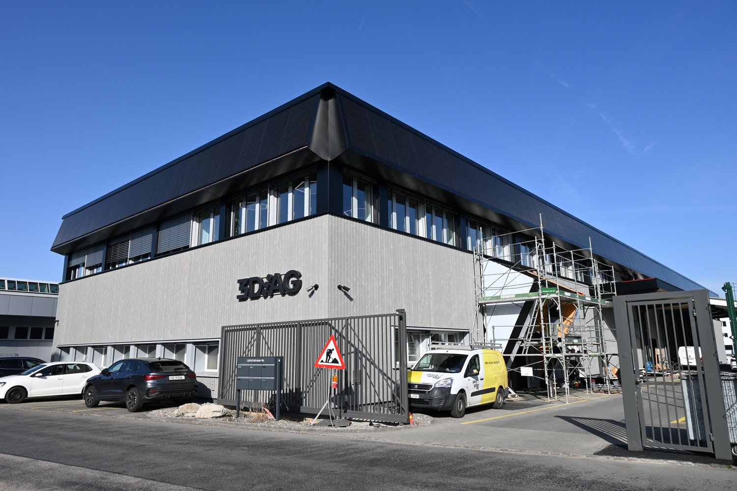 Office building, 3D/AG sign, multiple cars parked, two gates, construction scaffolding