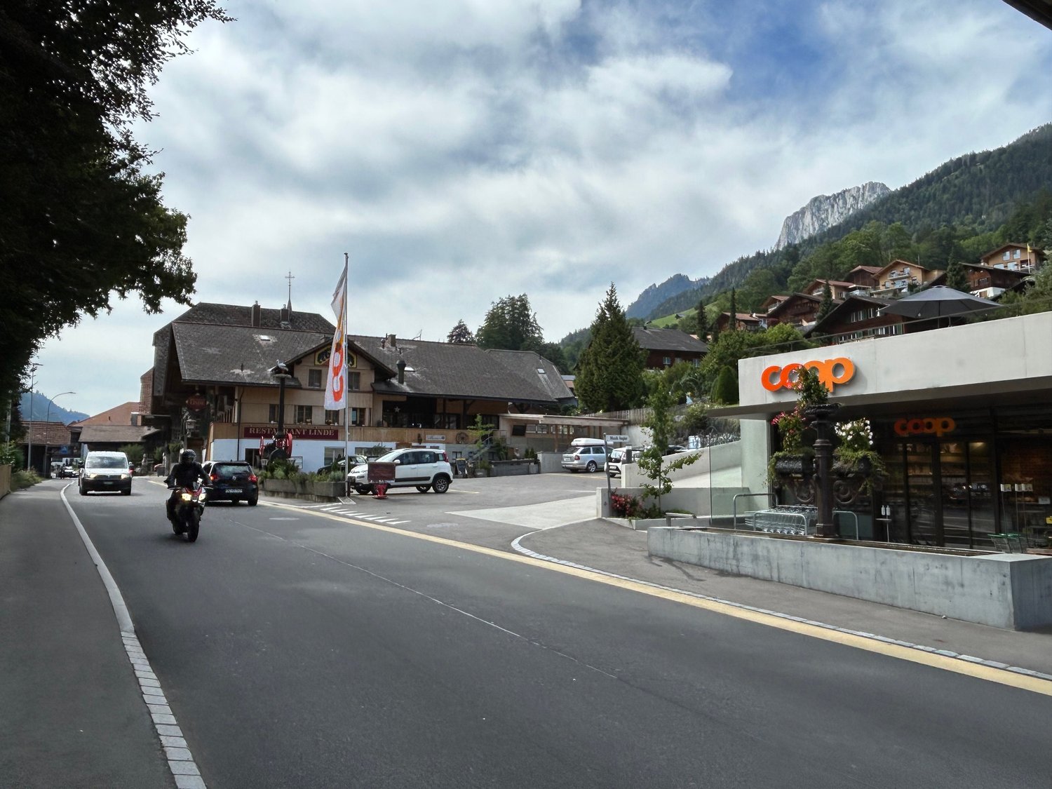 A street with a motorcycle and cars passing by, a building with a flag, a mountain in the background, and a shop with the name 'COOP'.