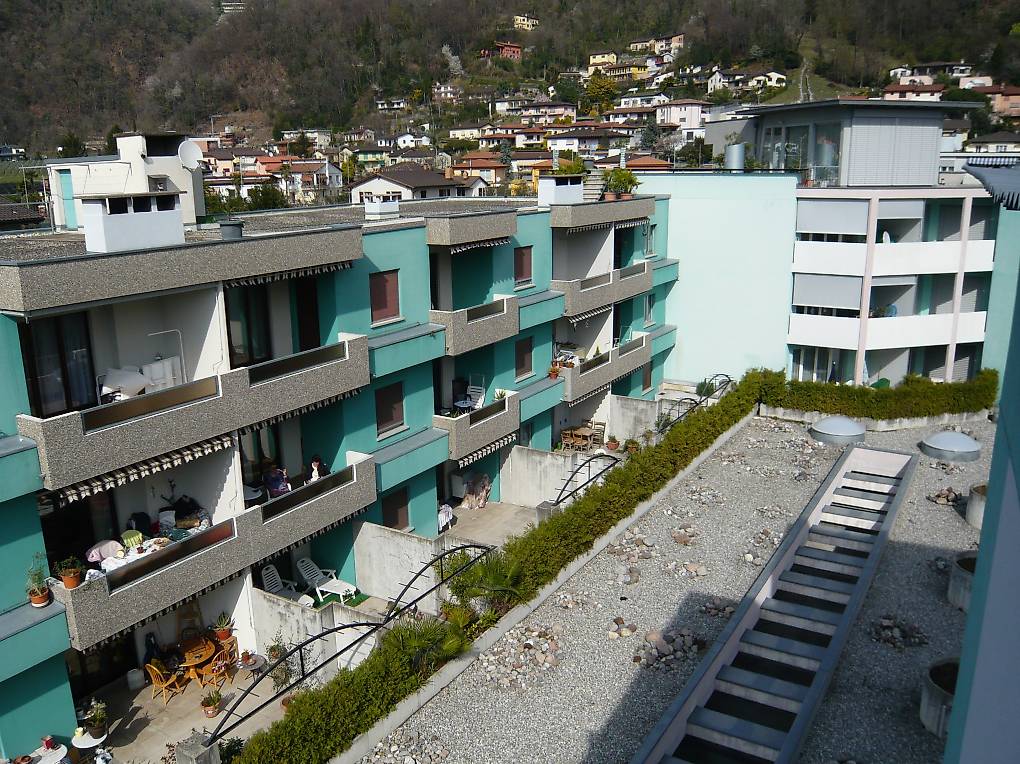 A multi-story building with multiple balconies, surrounded by greenery and a gravel pathway.