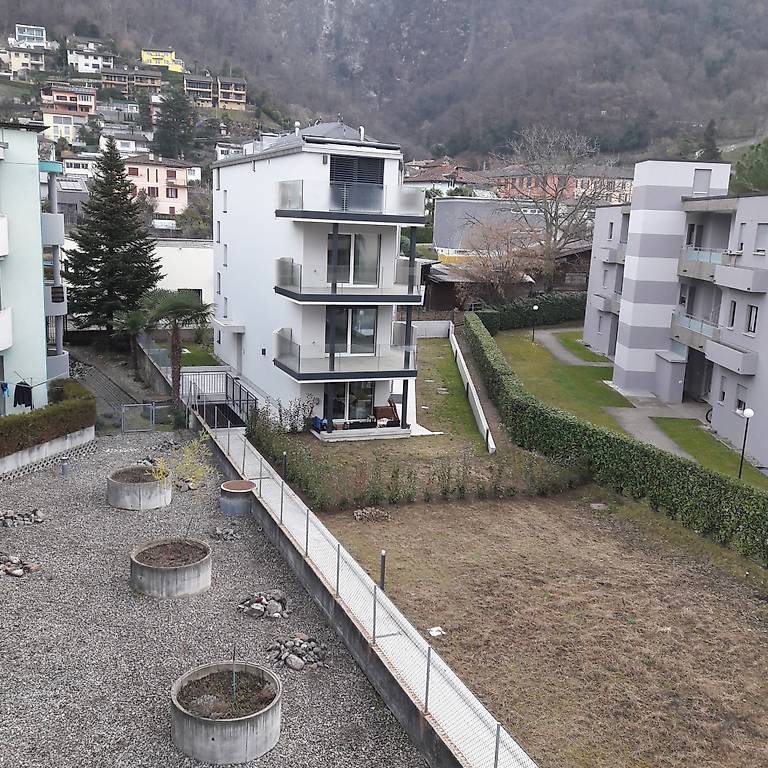 Aerial view of an apartment building in an urban setting with trees, a slope, and other buildings in the distance