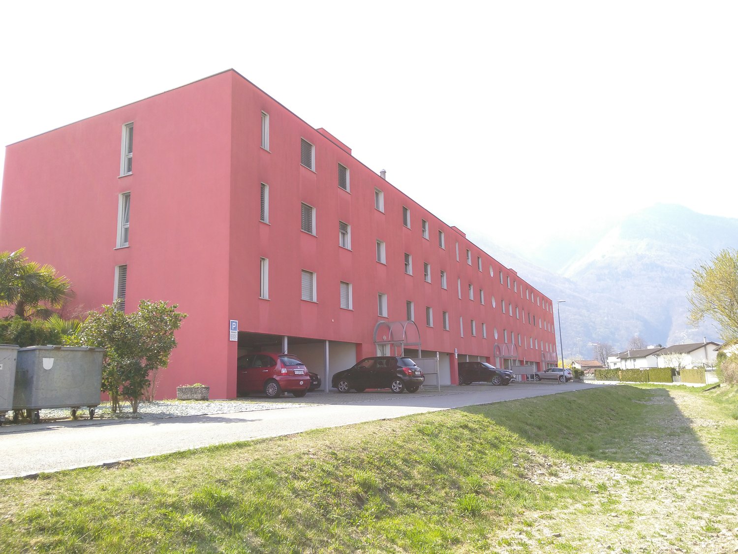 red building, multiple windows, parking lot, 3 cars parked, 2 trash bins, bushes
