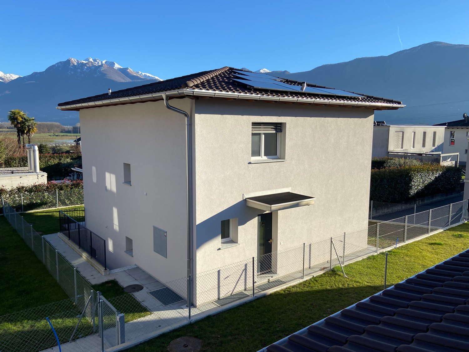 Modern 2 story house, solar panels on the roof, white exterior, fenced garden, mountains in the background