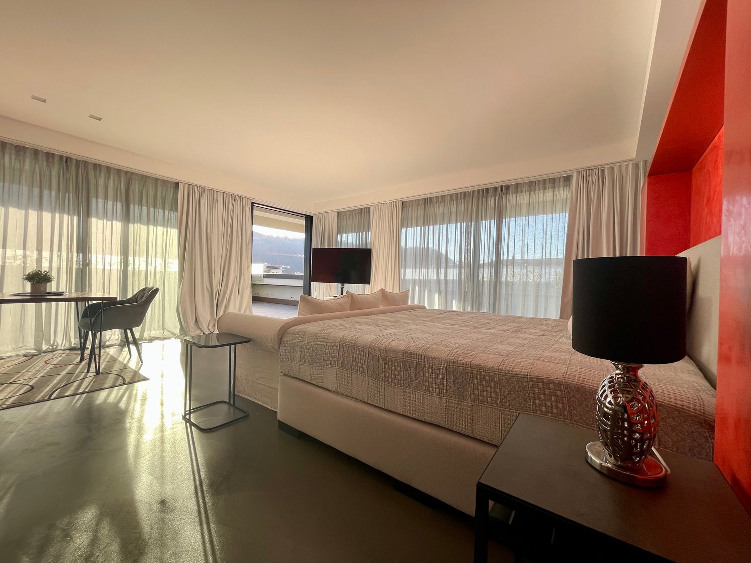 Modern and spacious bedroom with a king-size bed, large windows with white curtains, two armchairs, side table with lamp, small table with chairs, and a TV mounted on the wall. A rug is placed near the chairs.