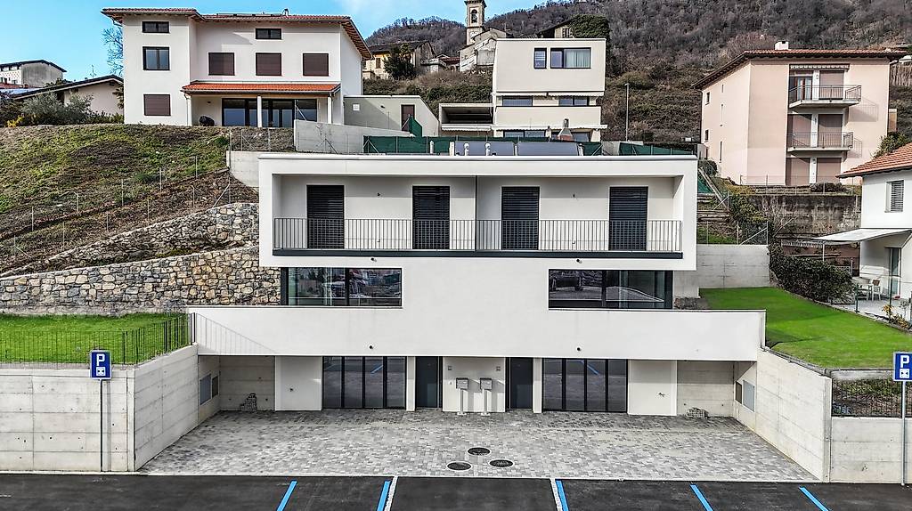Modern two-story house with a balcony, concrete wall, large garage, asphalt driveway, grassy areas, houses in the background, hilly surroundings, metal fences, and parking signs.