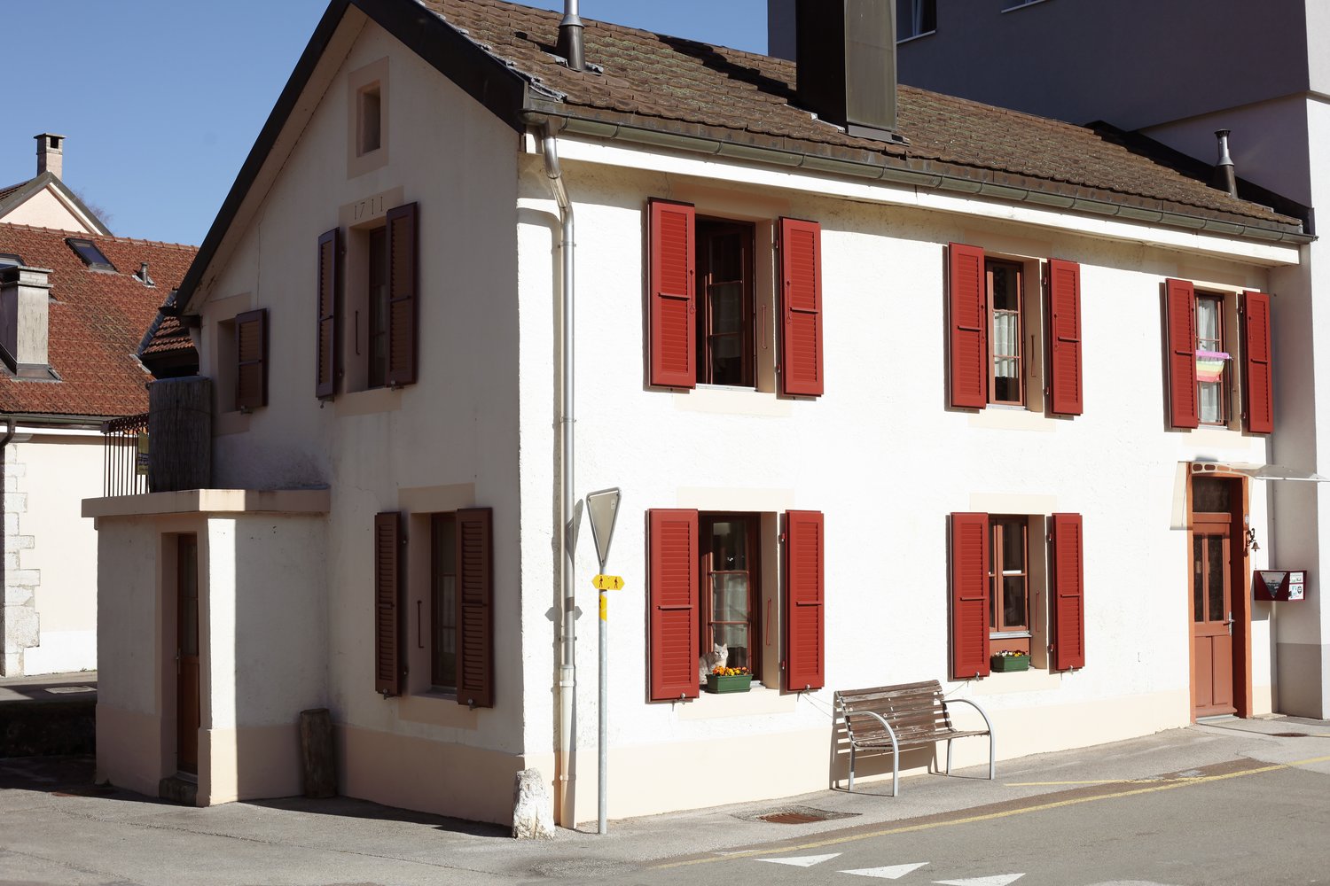 white house with red shutters, multiple windows, brown door, bench on the sidewalk