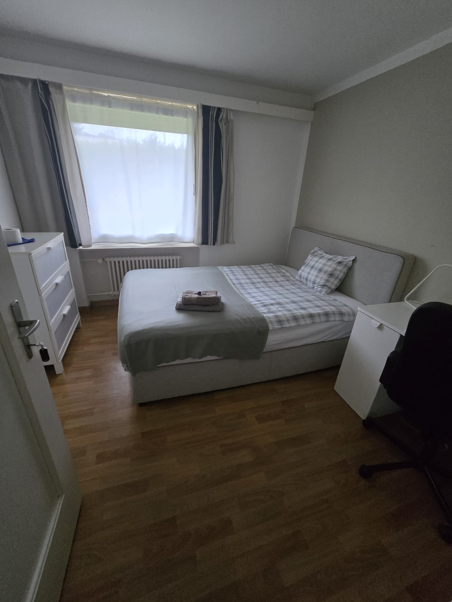 A bedroom with a single bed, a pillow, and a blanket. It has a small window with blue curtains and a white radiator below. On the left, there is a white chest of drawers. On the right, there is a small desk and a chair. The floor is wooden.