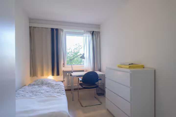 small bedroom with bed, chair, desk with lamp, and drawers