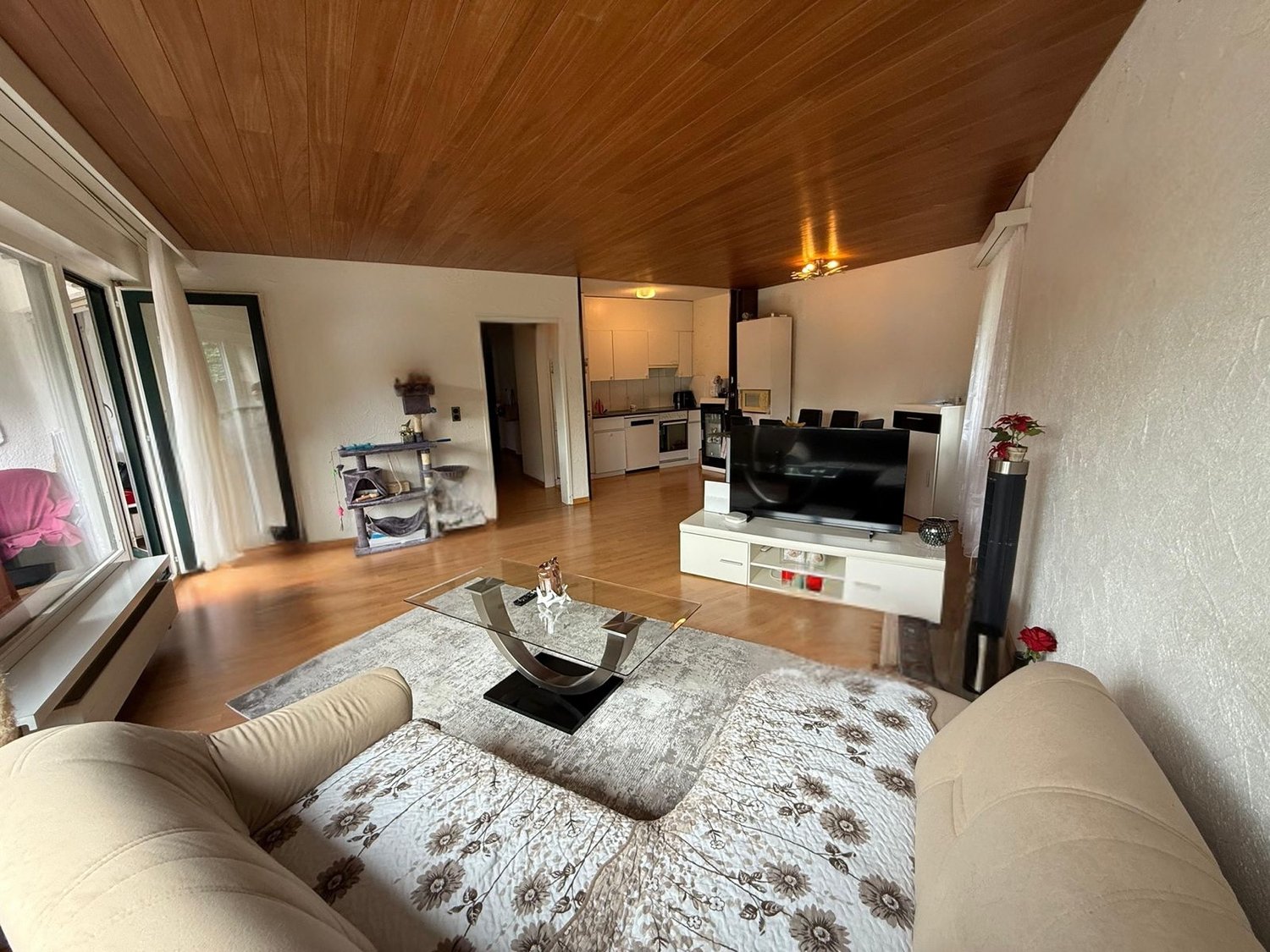 Open living room with hardwood floors, beige couches, glass center table, large windows, and a TV stand with a flatscreen TV. There is a doorway leading to the kitchen.