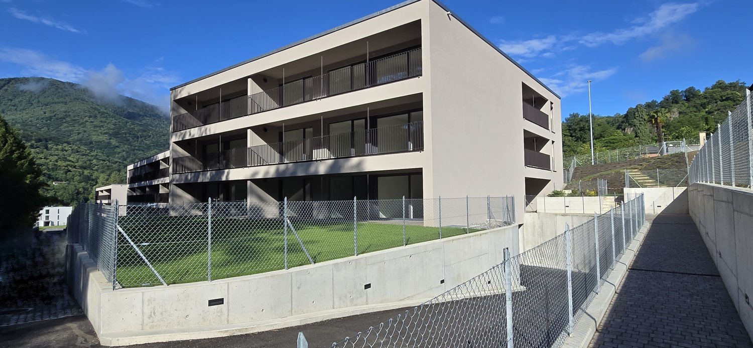 Modern apartment building, multiple floors, balconies, concrete walls, fenced courtyard, green grass