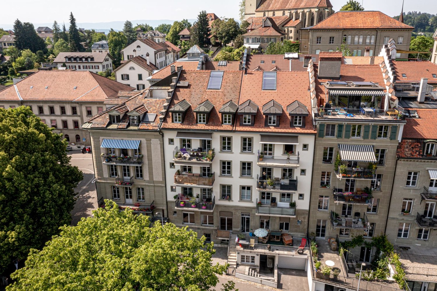 This image shows a panoramic view of a historic European city, with a mix of traditional and modern architecture. The buildings have red tiled roofs, and many have balconies or terraces visible. There are trees and greenery throughout the cityscape, creat