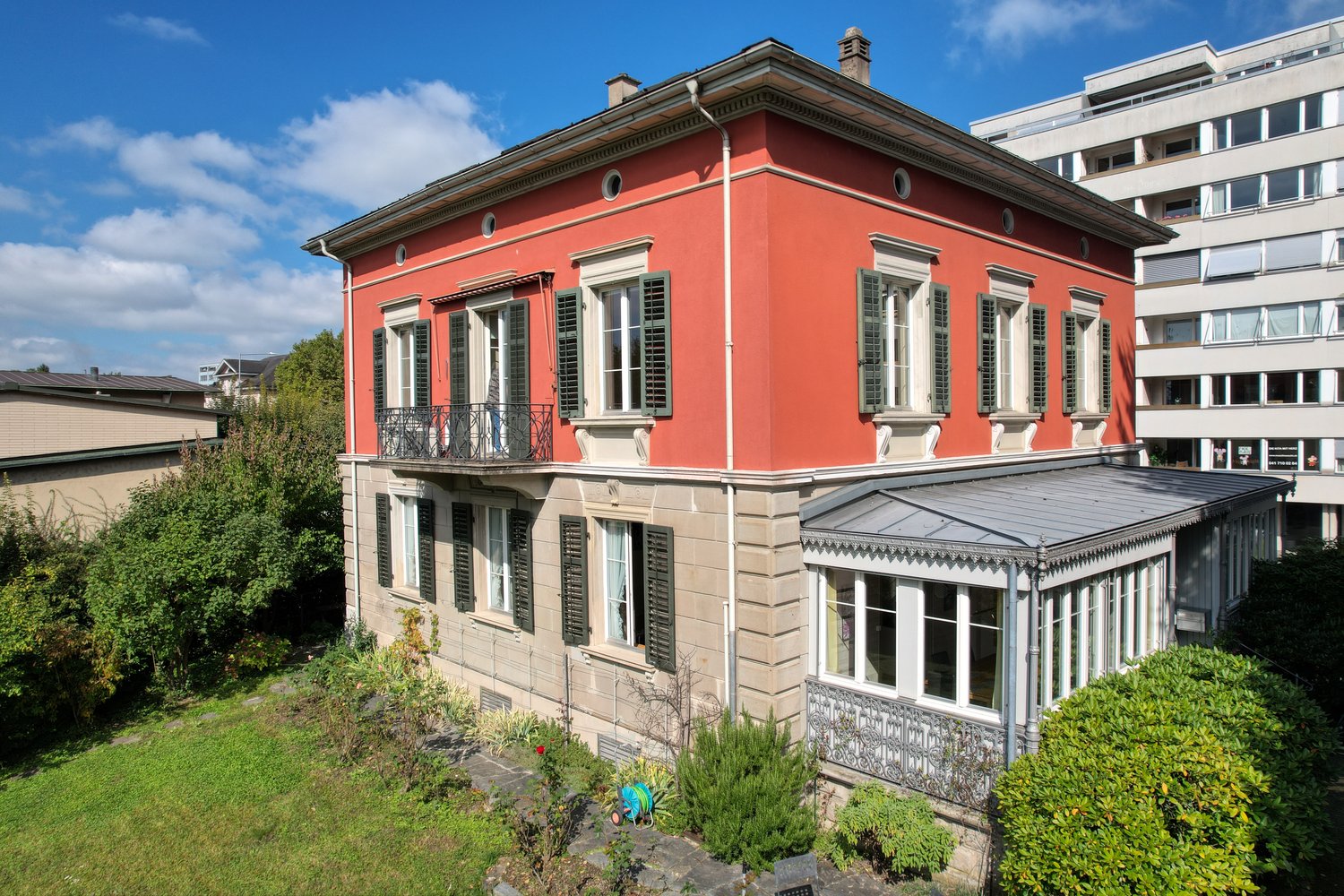 Large villa, red paint, multiple windows, 2 floors, balcony, small porch, green trees and bushes