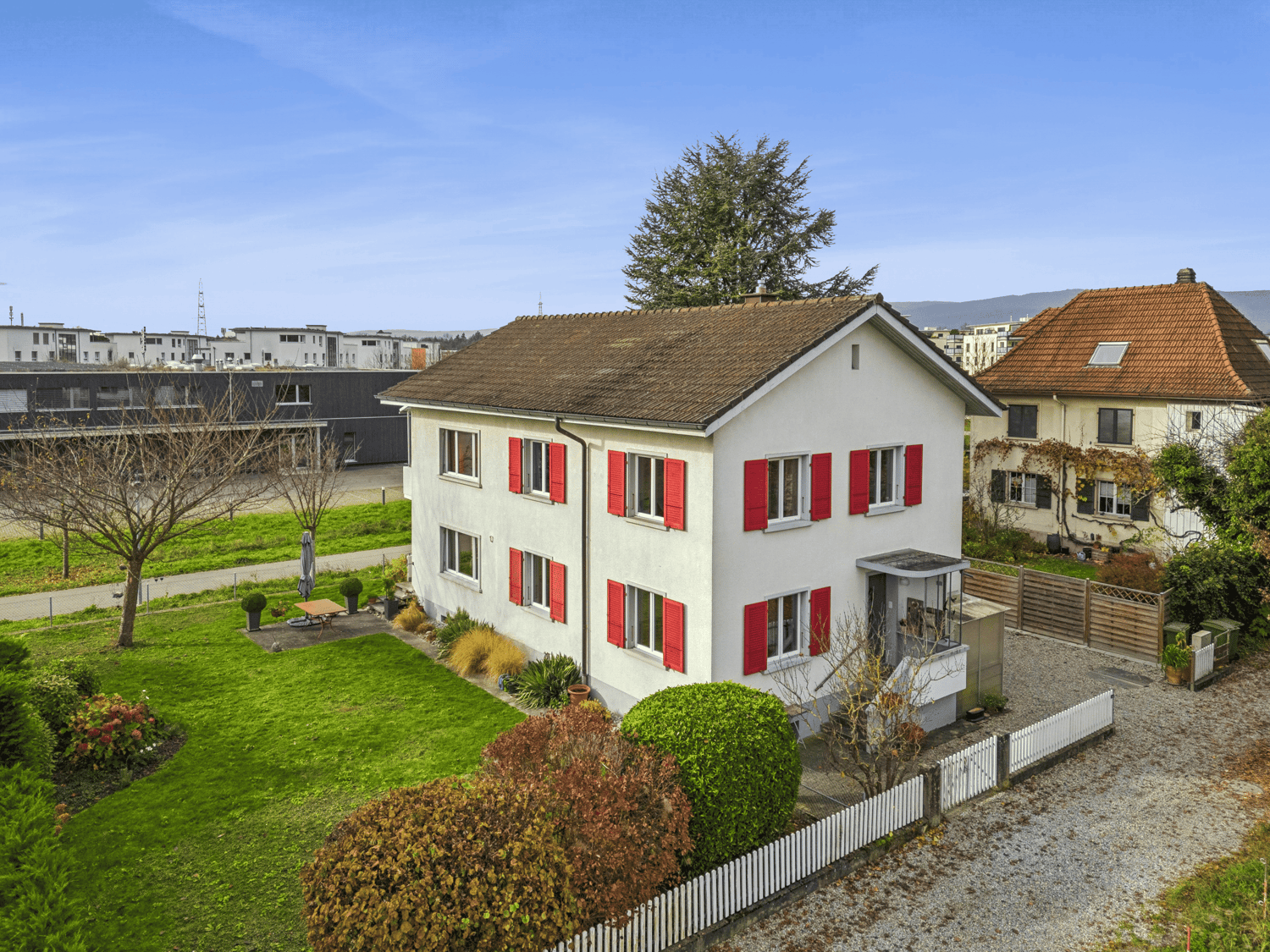 Detached house with brown roof, white walls, red shutters, fenced garden, gravel driveway, white fence