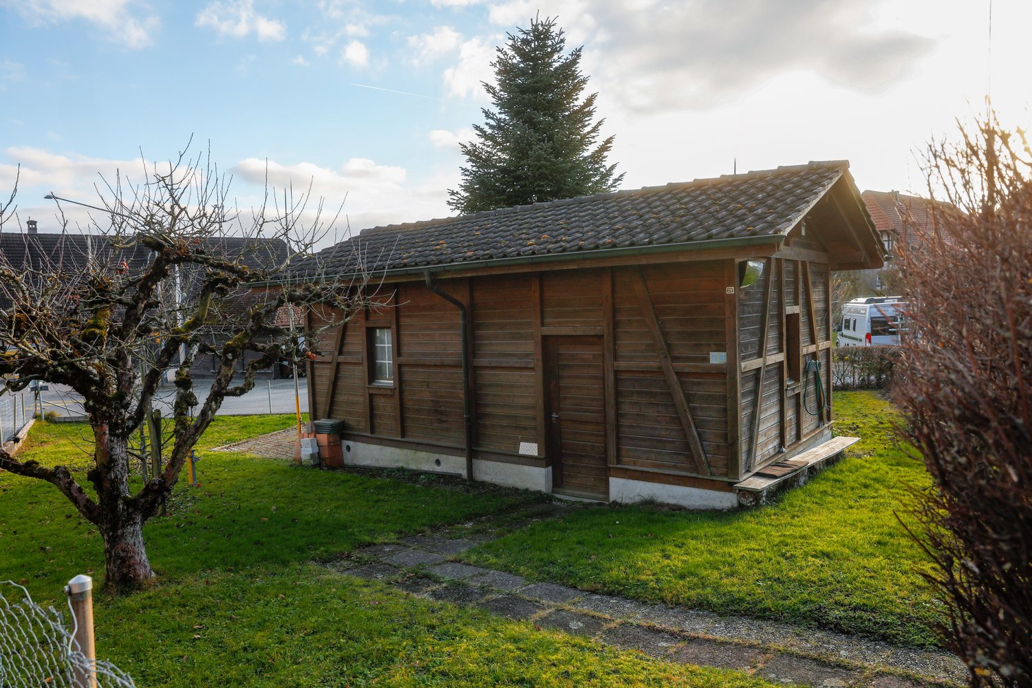 small wooden house, single-storey, slanted roof, windows, wooden entrance, pathway, tree, grass, car parked