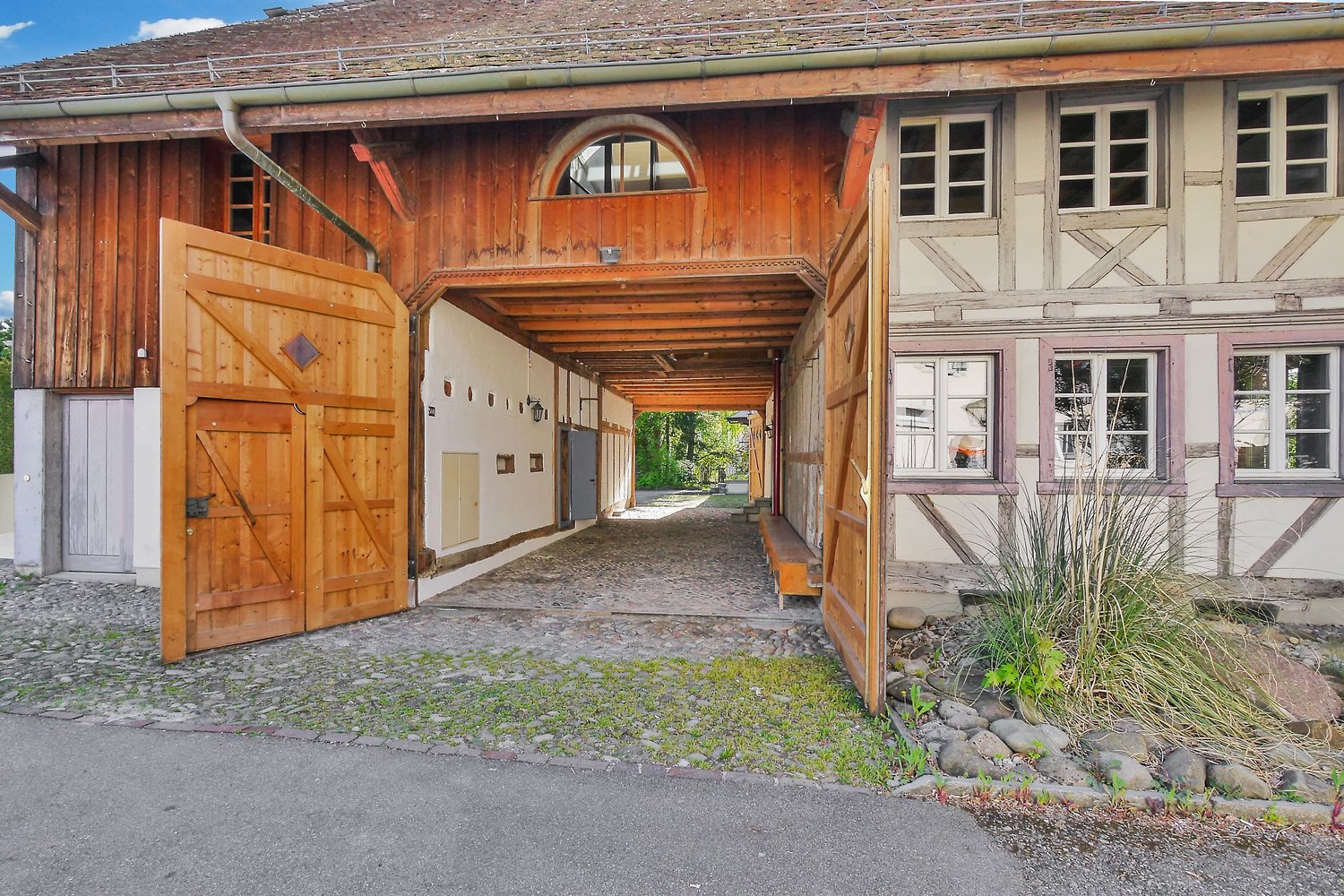 Exterior view of a wooden and brick house with large wooden double doors, small wooden bench in the courtyard, driveway, paved front courtyard