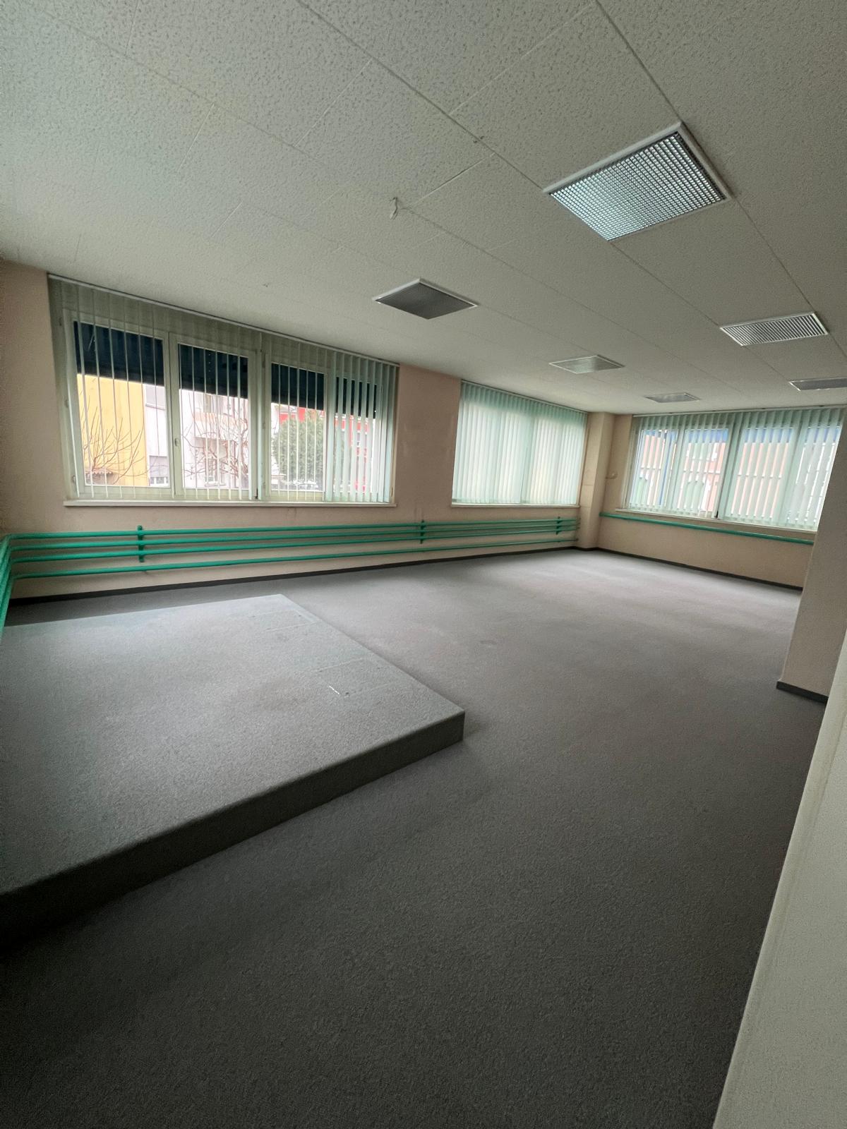 An empty room with gray carpet floor, white ceiling, large windows with metal blinds, and an elevated platform on the left.