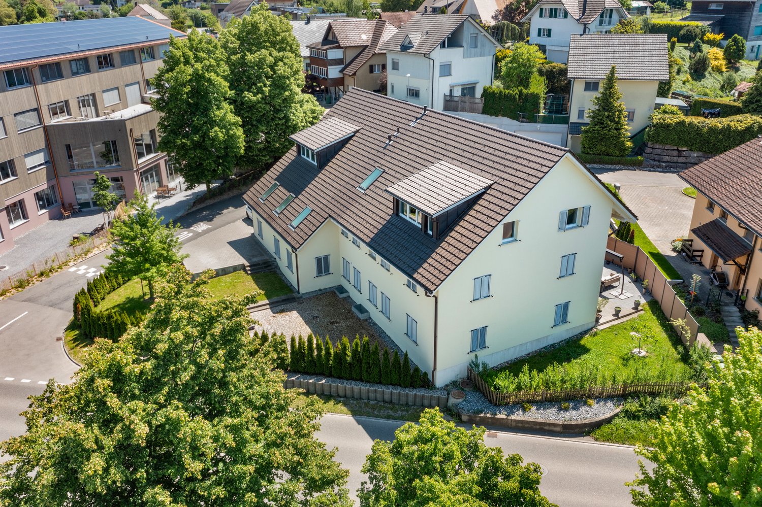 3-story house with brown roof, white walls, solar panels on neighbor's building, surrounded by trees and other houses