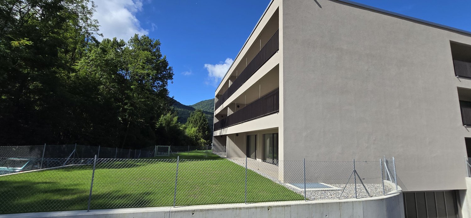 Modern apartment building, gray exterior, balconies, fenced yard, green grass, trees