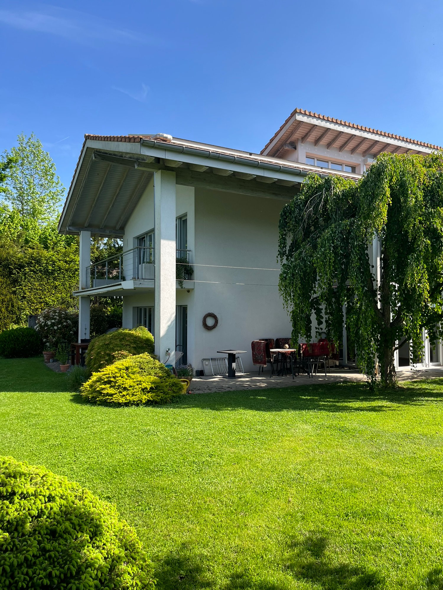 modern house with 2 floors, red tiled roof, large balcony on 2nd floor, garden with lawn and trees, outdoor dining area