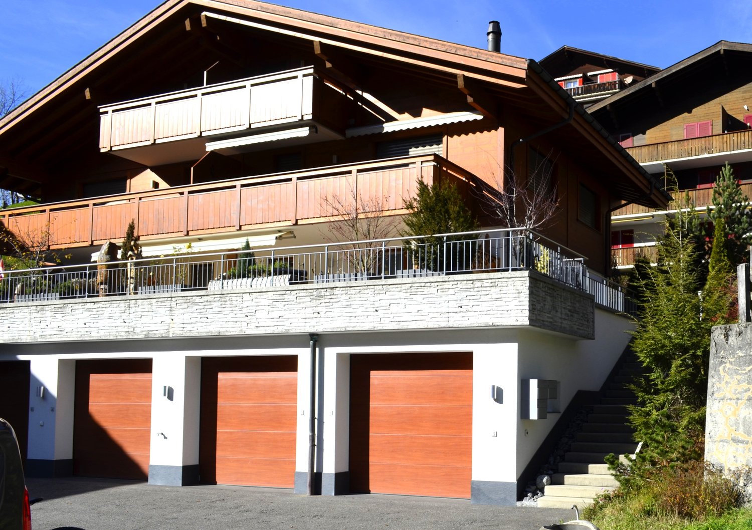 A two-story chalet-style house with a brown exterior and white accents, featuring three wooden garage doors and a staircase leading to the upper floor.
